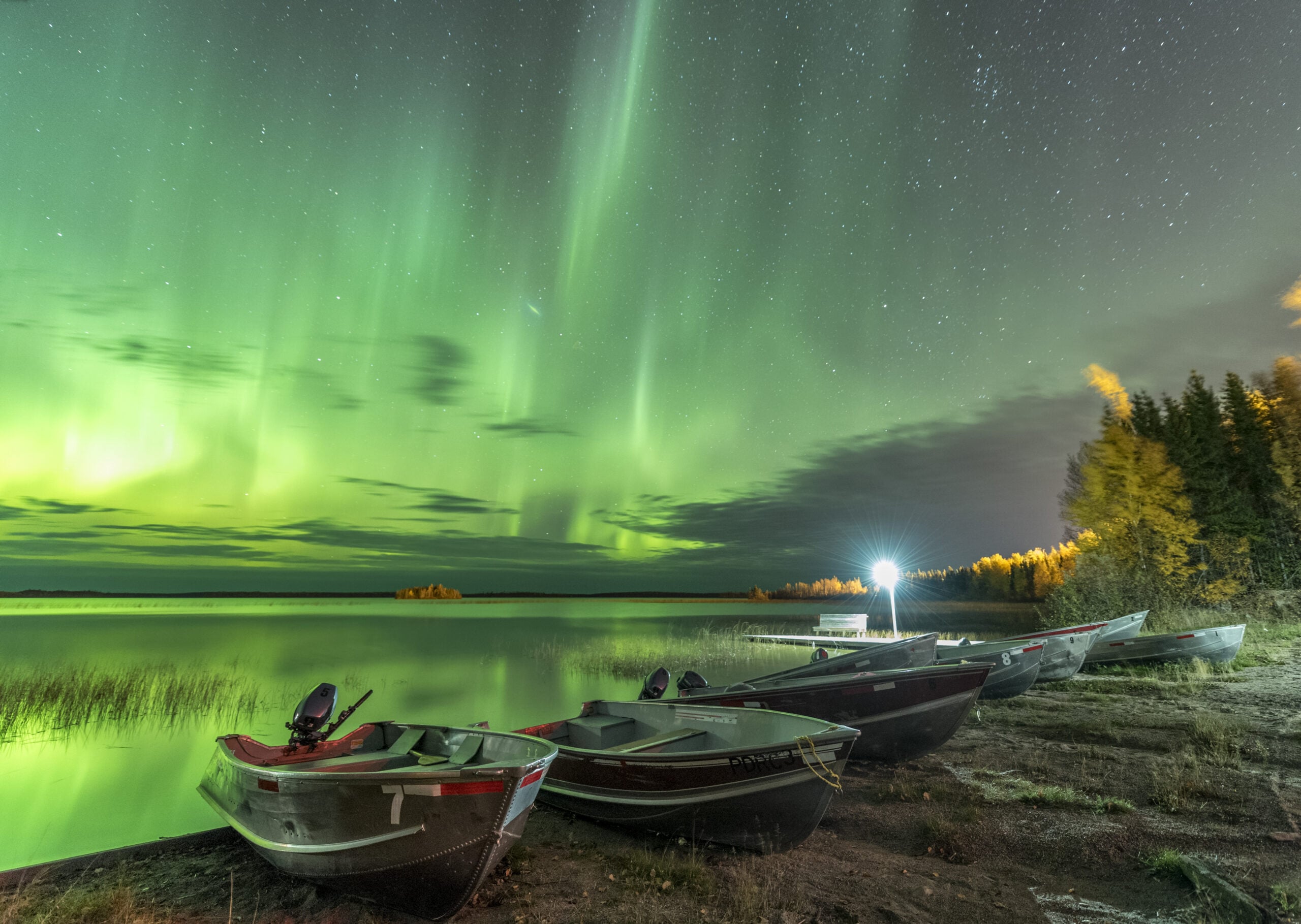 Several boats rest on a shore near calm water, under a vivid green aurora borealis in the night sky, with trees and a bright light in the background.
