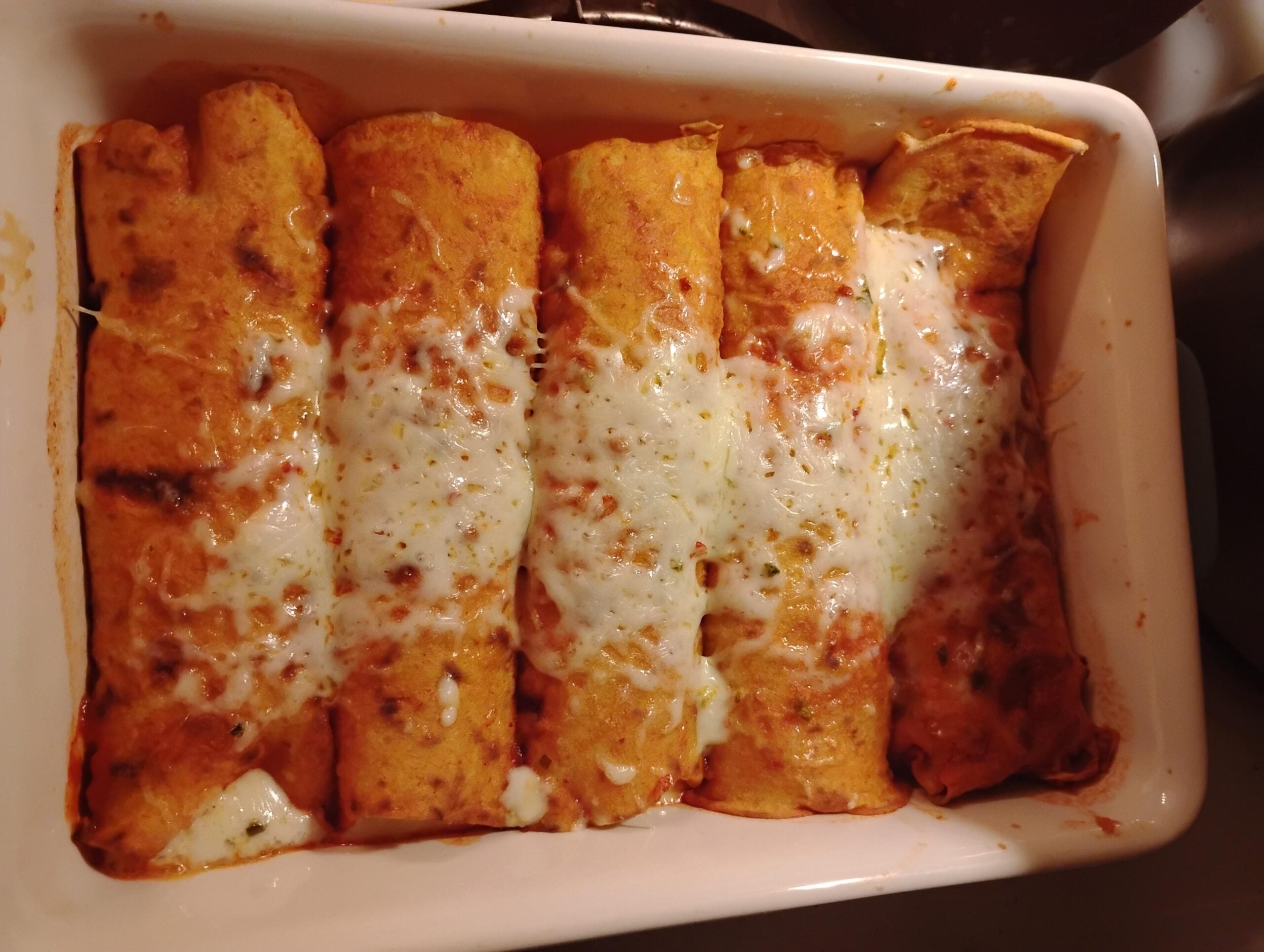 Five baked enchiladas in a white dish, topped with melted cheese and red sauce. The edges are slightly browned and the cheese is bubbly, giving a warm and appetizing appearance.