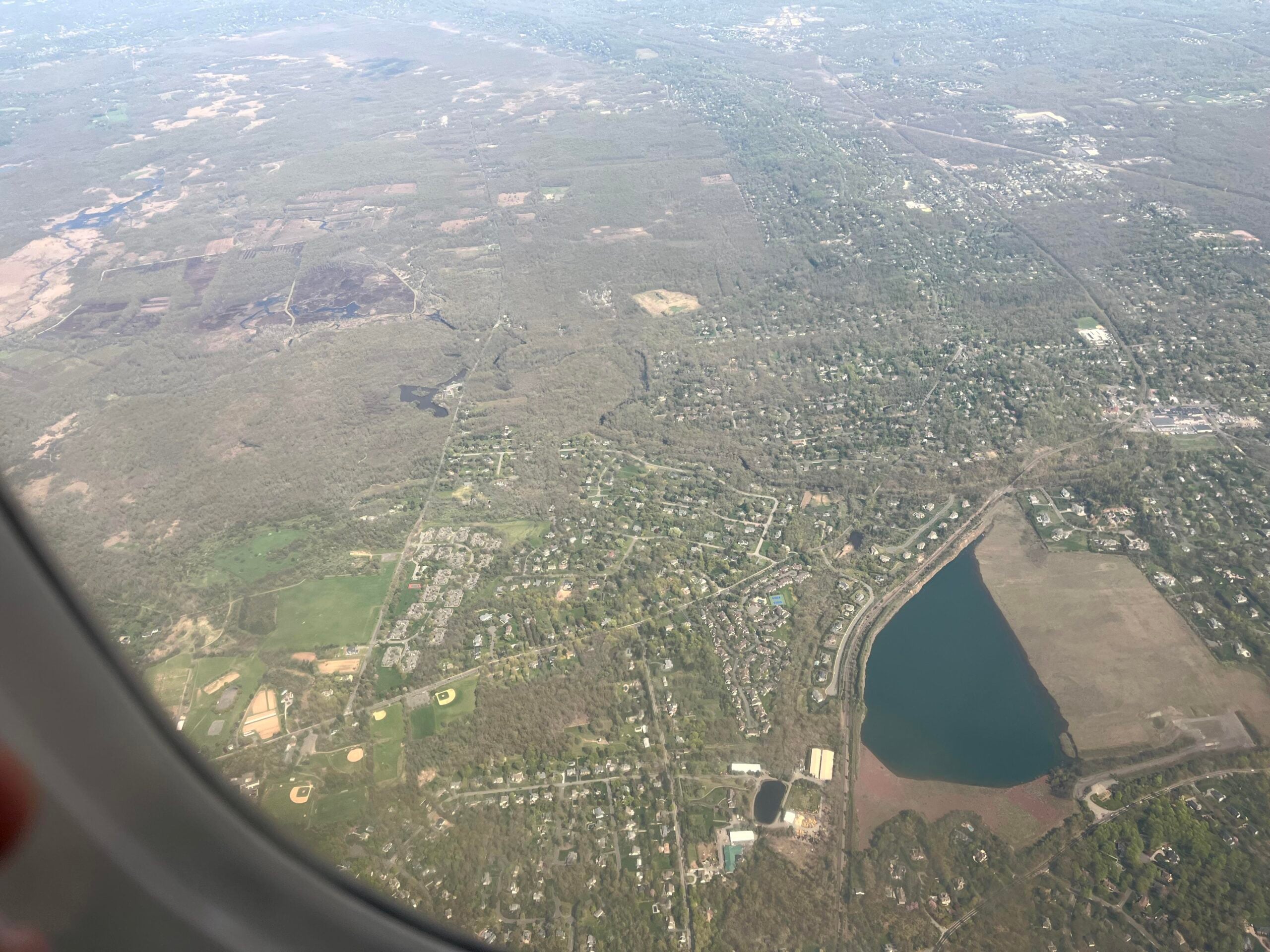 Aerial view from airplane windows showing a patchwork of suburban houses, roads, a large lake, and a mix of forested and open land.
