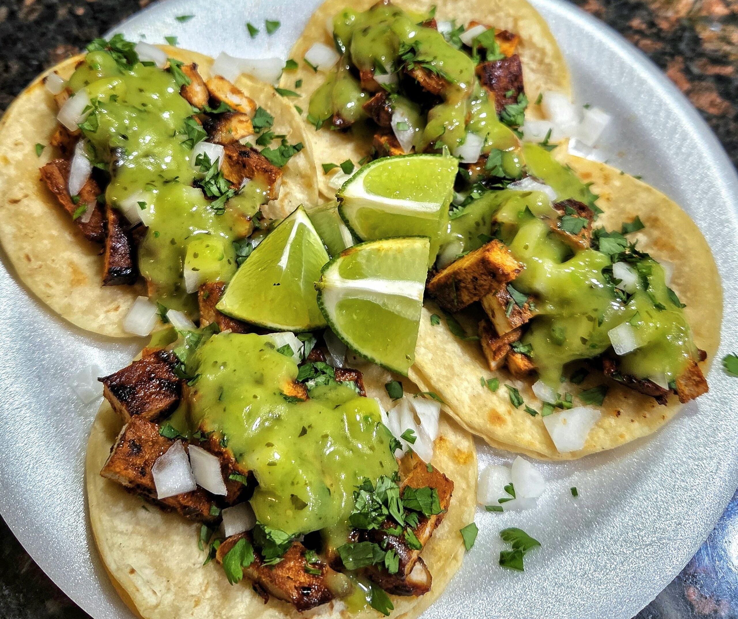 Four tacos filled with grilled meat, diced onions, cilantro, and topped with green salsa are served on a white plate with lime wedges in the center.