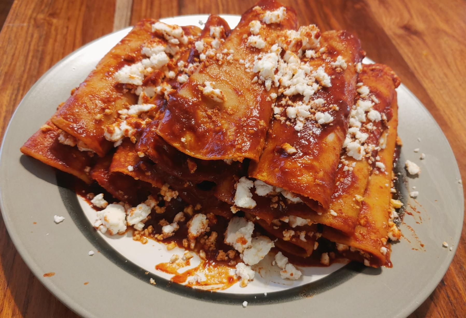 A plate of red enchiladas topped with crumbled cheese, served on a gray-rimmed plate atop a wooden table.
