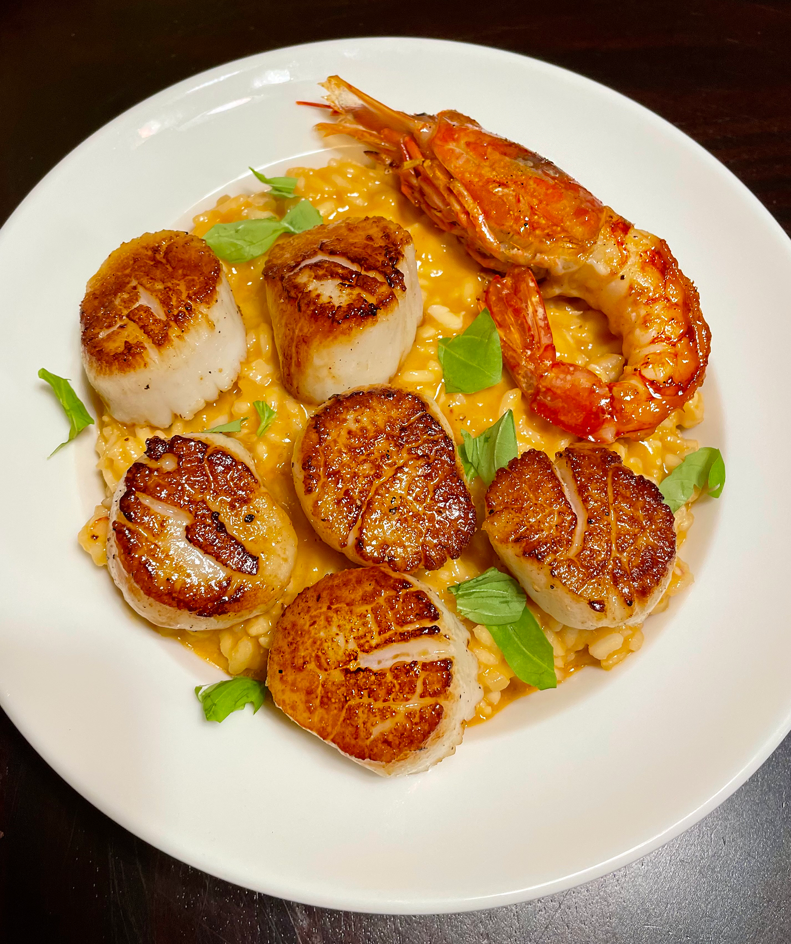 A white plate with six seared scallops, a large grilled prawn, and fresh green herbs on creamy risotto.