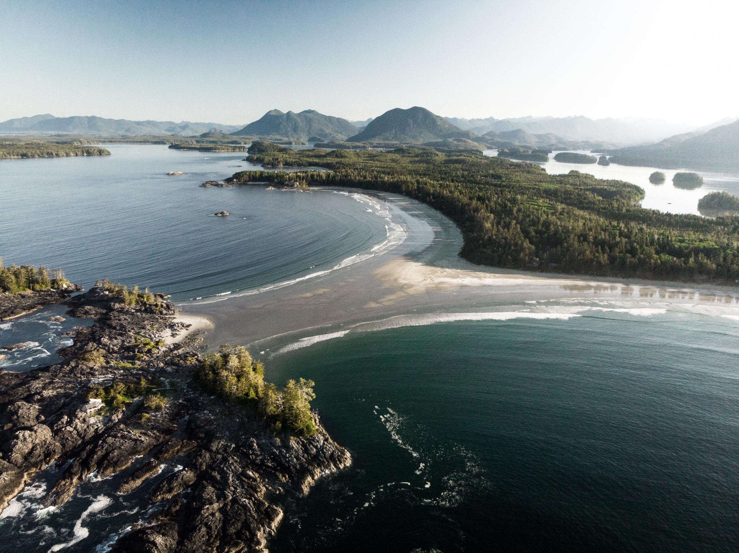 Aerial view of a coastal landscape with curved sandy beaches, forested islands, and gentle waves meeting the shore under a clear sky, with distant mountains in the background.