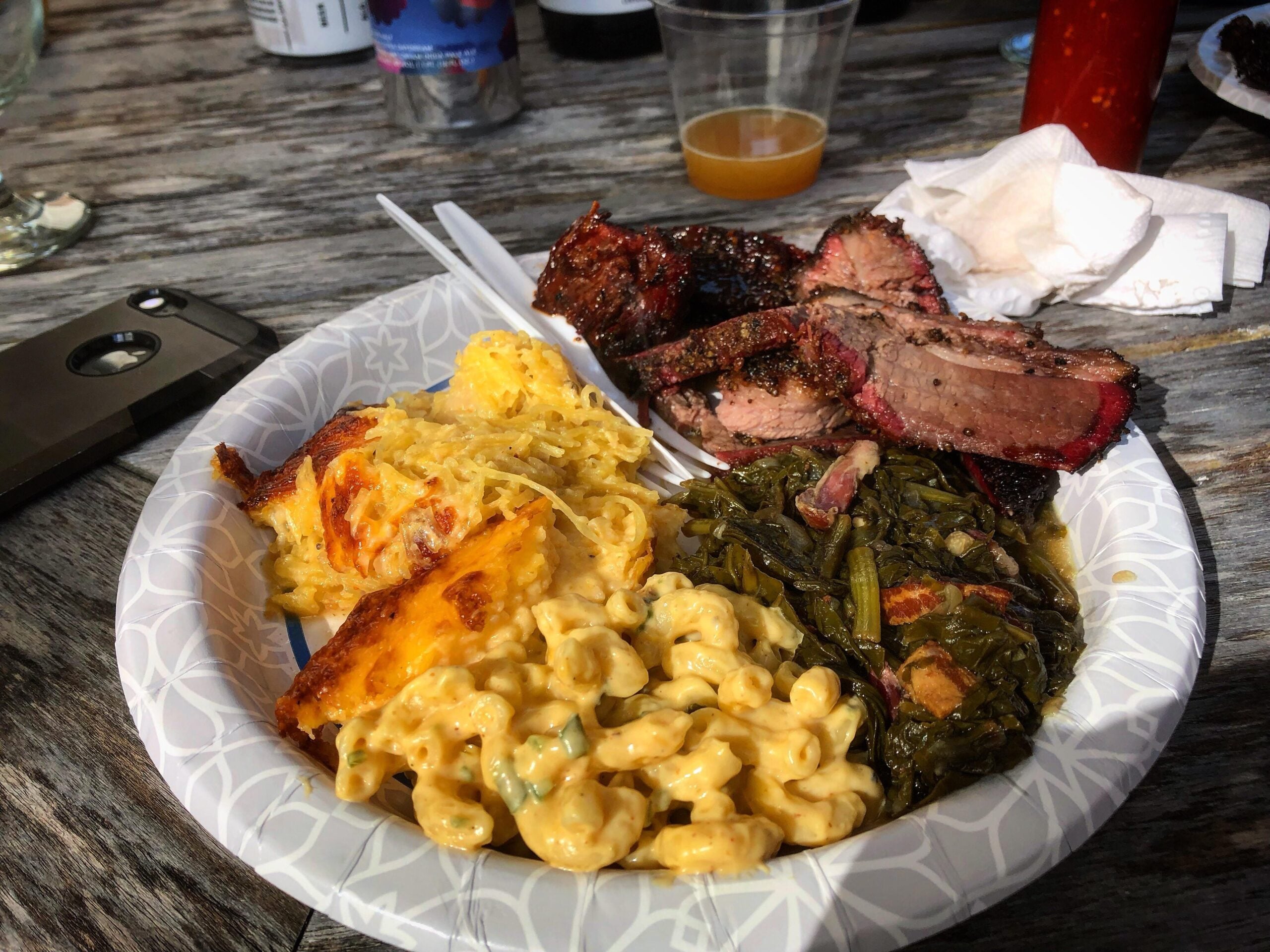 A paper plate with macaroni and cheese, collard greens, cheesy casserole, and slices of smoked brisket sits on a wooden table with drinks, napkins, and a phone nearby.