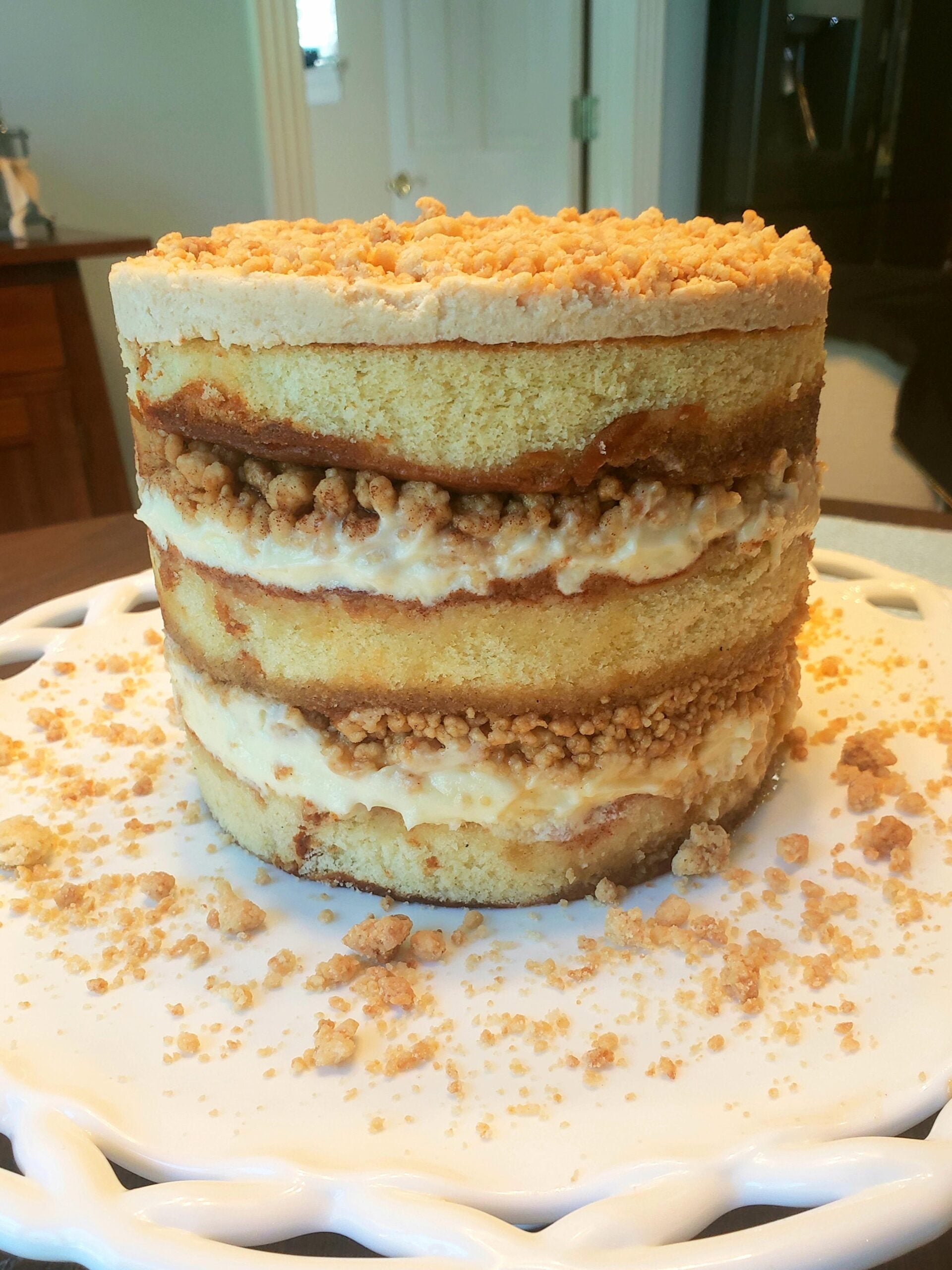 A tall, naked-layer cake with three layers of yellow sponge, creamy filling, and crumb topping, displayed on a white cake stand with crumbs scattered around.