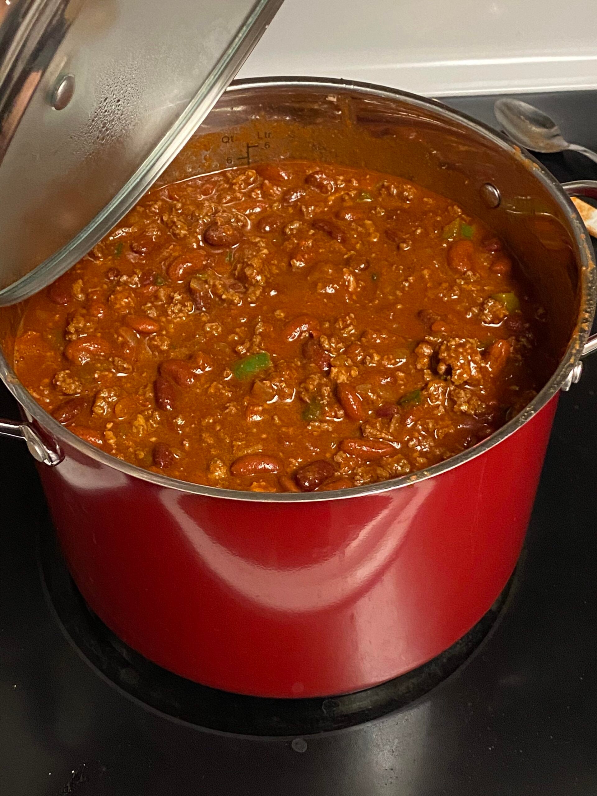A red pot of chili with beans, ground meat, and tomatoes simmering on a stovetop, partially covered with a lid.