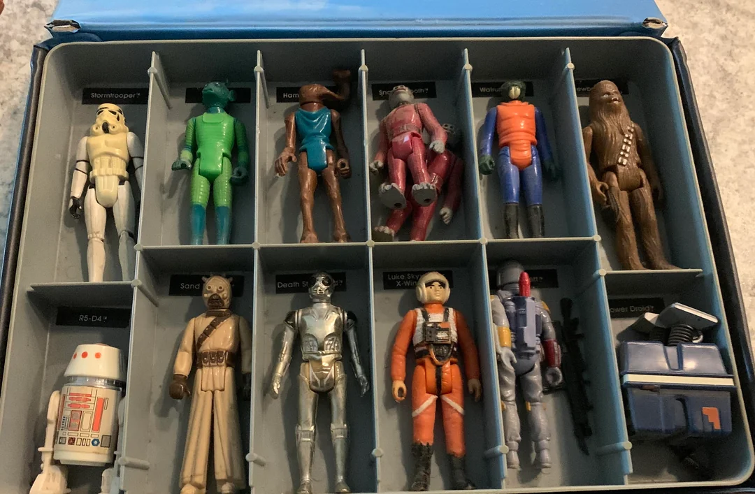 A blue case with vintage Star Wars action figures, including a Stormtrooper, Han Solo, Chewbacca, and droids, each in separate compartments. The box is open on a gray surface.