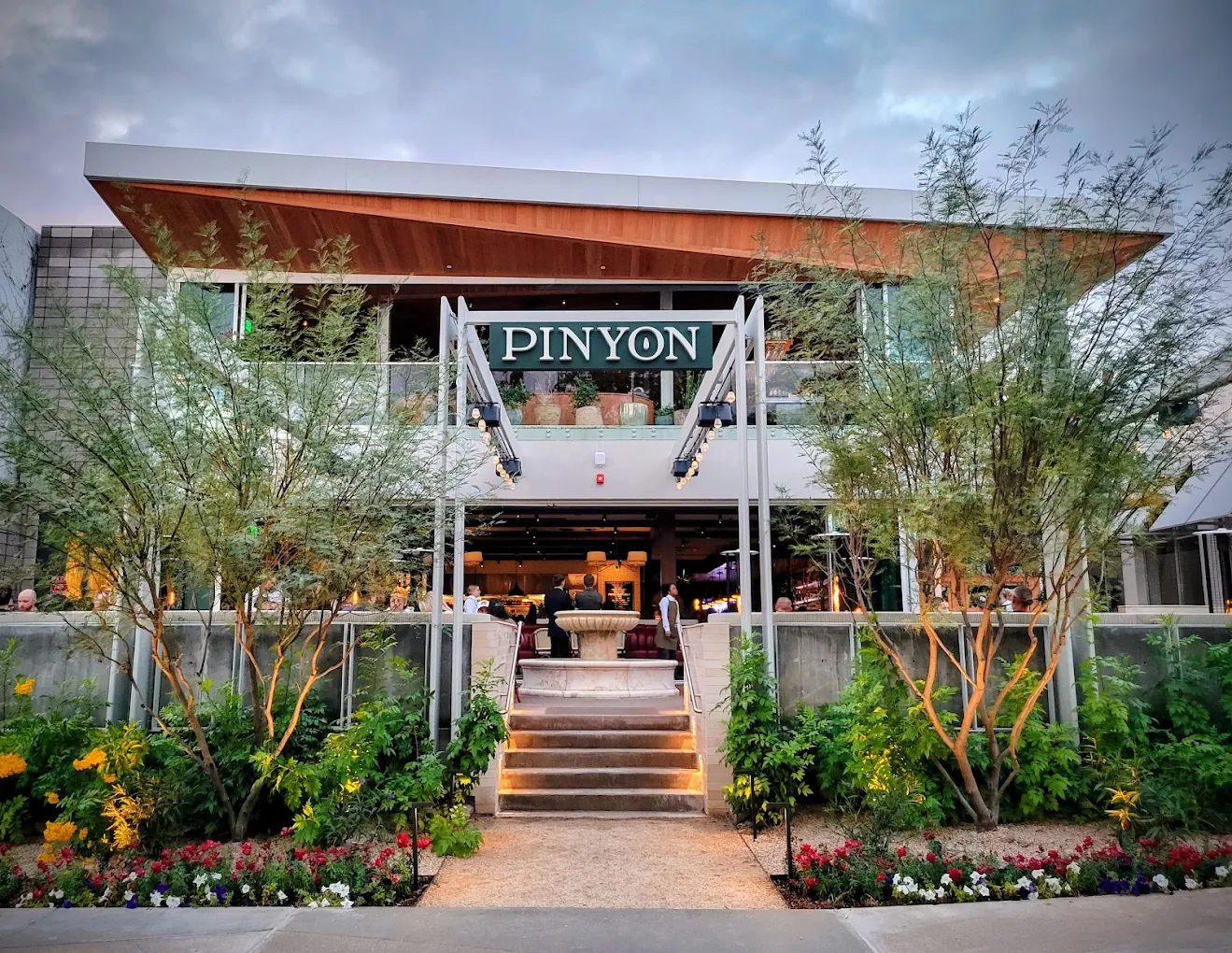 A modern restaurant named PINYON with large windows, outdoor seating, and lush landscaping. Steps with lights lead to the entrance, surrounded by green plants and flowers. Best-rated Mediterranean restaurants.