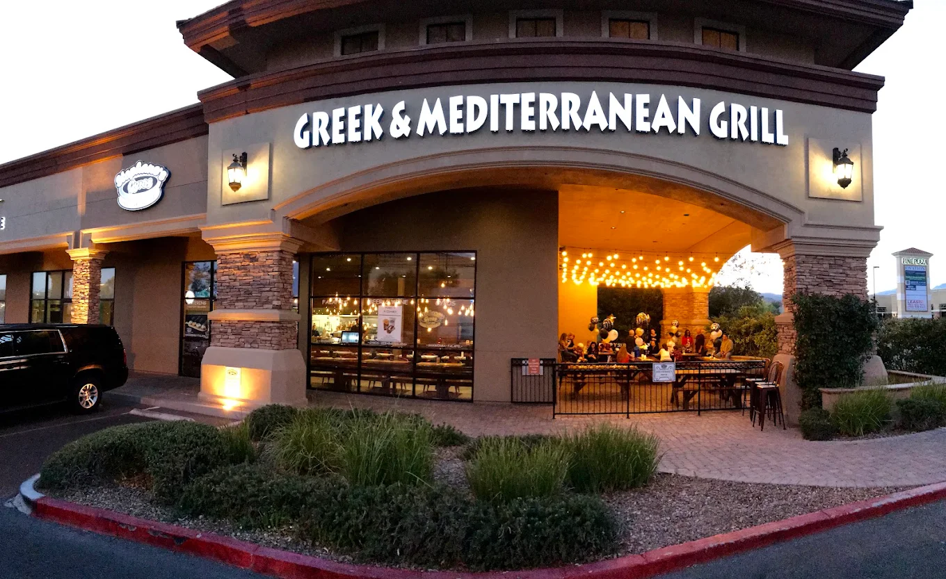 A Mediterranean restaurant exterior at dusk, with large windows showing warmly lit diners inside and some people seated on the outdoor patio. The sign reads “Greek & Mediterranean Grill.”