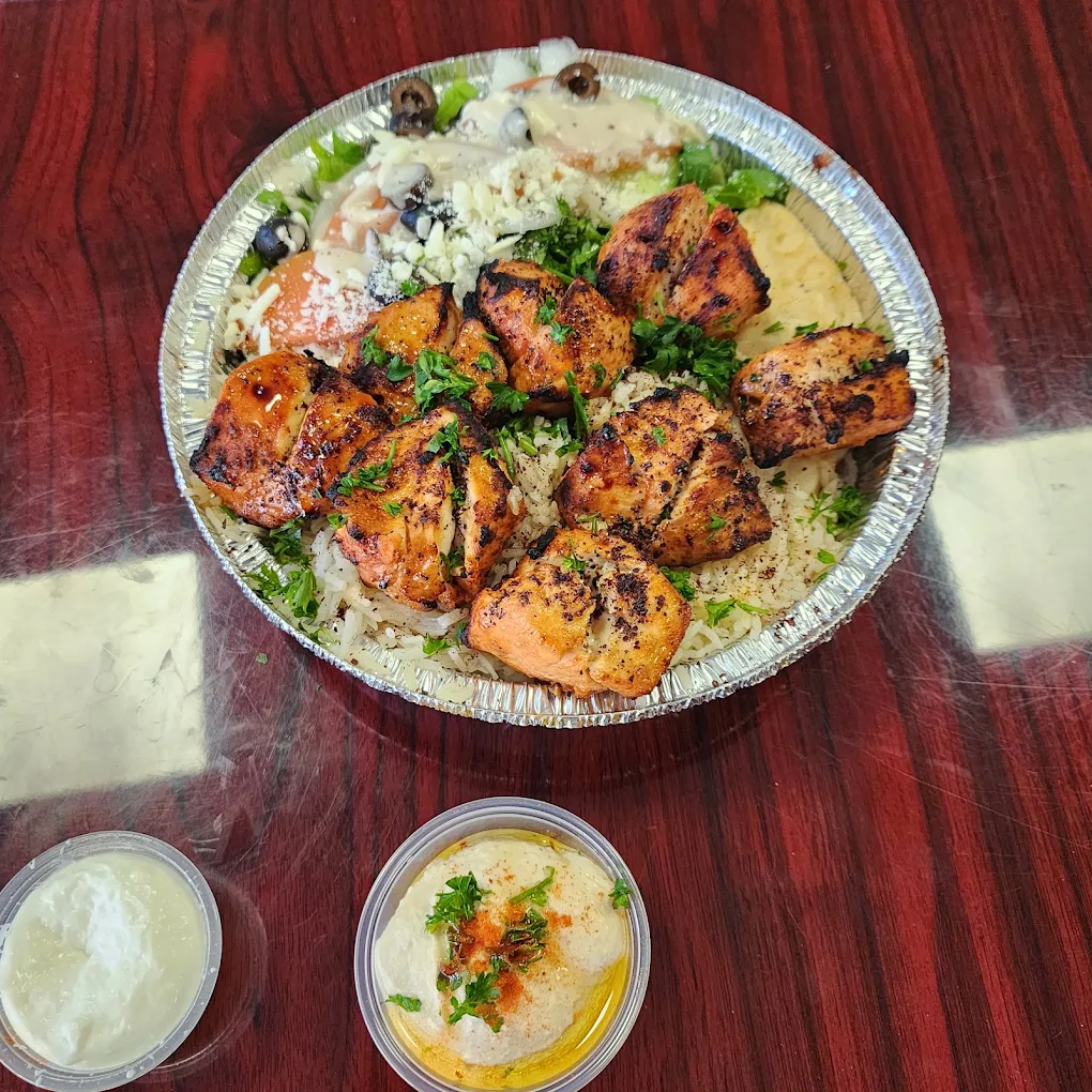 A foil container with grilled chicken over rice, salad, and olives, served with a side of creamy white sauce and hummus, on a wooden table.