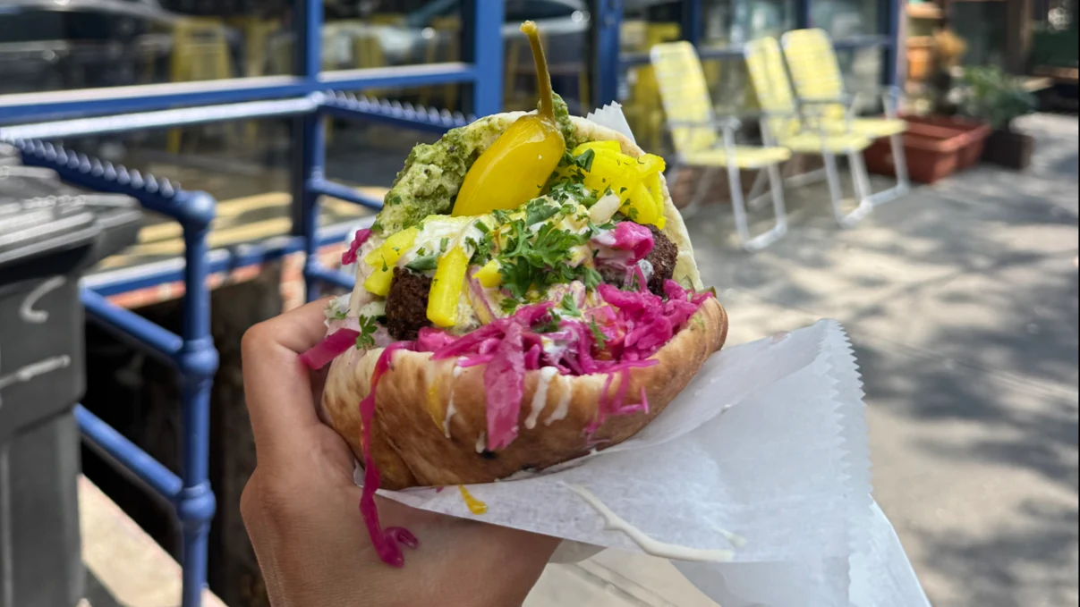 A hand holds a pita stuffed with falafel, yellow pickles, red cabbage, herbs, and creamy sauce. Outdoor seating with yellow chairs and tables is visible in the blurred background.