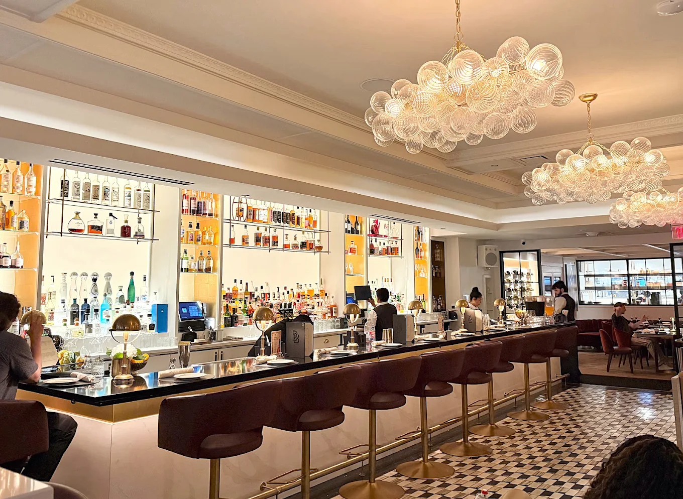 A modern bar with high-backed chairs, glass shelves displaying bottles, and globe chandeliers. Bartenders work behind the counter, and diners sit at tables in the background. The floor features a black-and-white tile pattern.