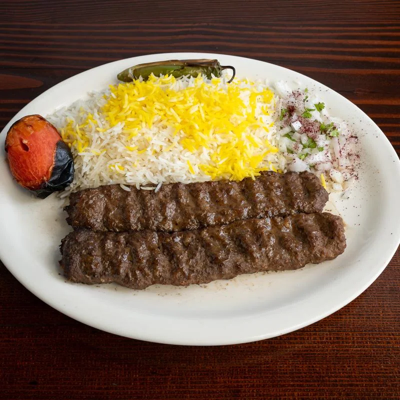 A plate with two grilled kebabs, saffron rice, a roasted tomato, a grilled pepper, and a small side of chopped onions with herbs on a dark wooden table.