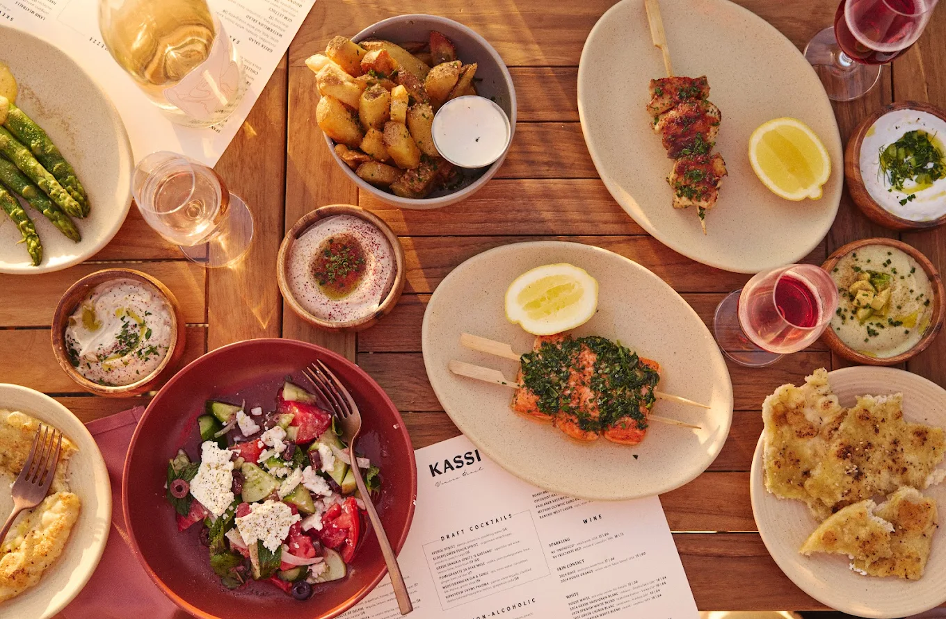 A table set with Mediterranean dishes, including grilled skewers, potatoes with dip, Greek salad, flatbread, drinks, and menus on a wooden surface in warm lighting.