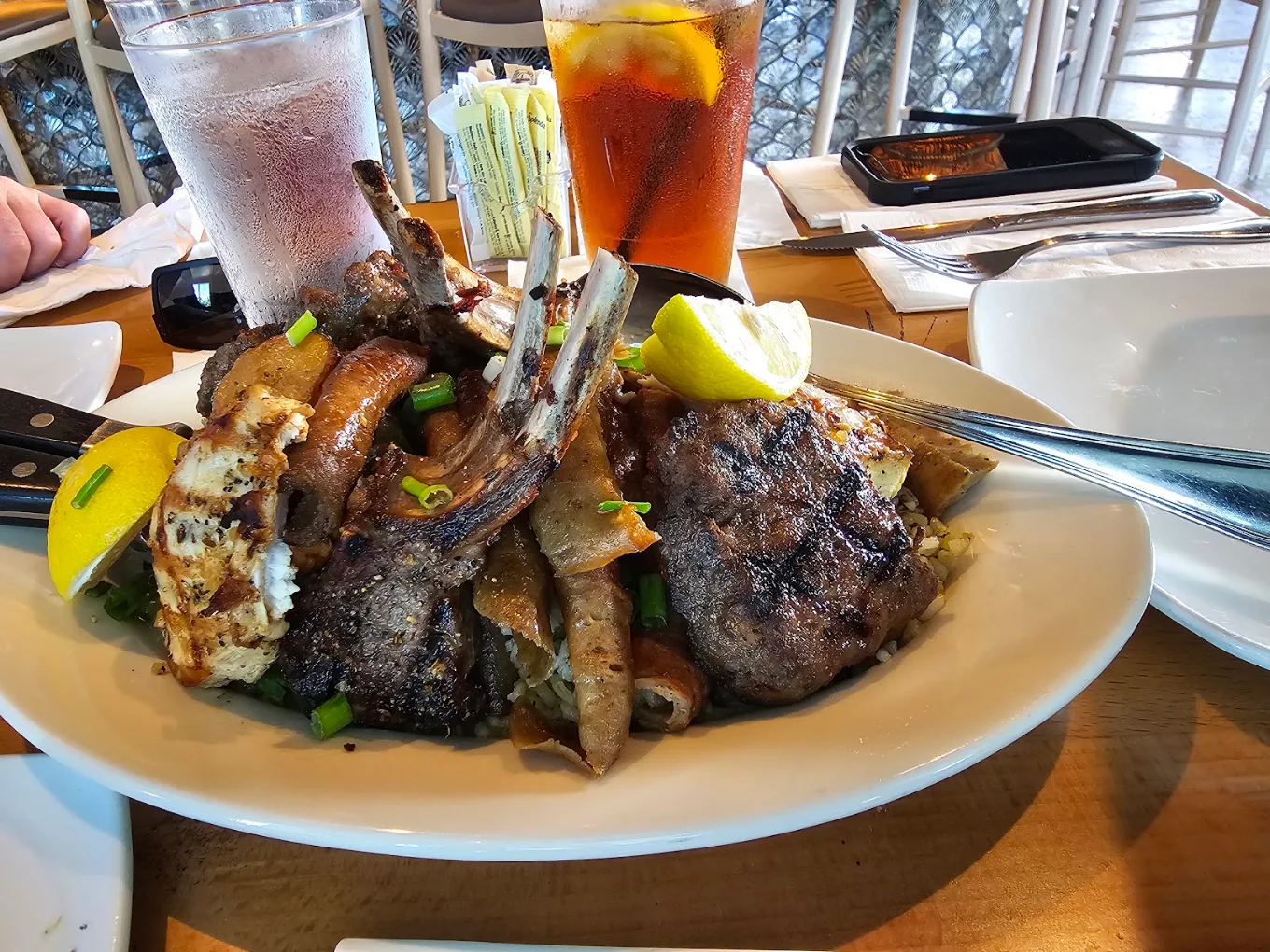 A white plate filled with assorted grilled meats, garnished with lemon wedges and green onions, sits on a wooden table next to drinks and utensils.