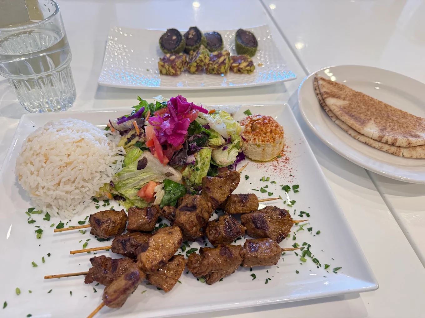A plate with grilled meat skewers, white rice, salad, and a scoop of hummus. In the background, grape leaves stuffed with rice on another plate, pita bread on a side plate, and a glass of water.