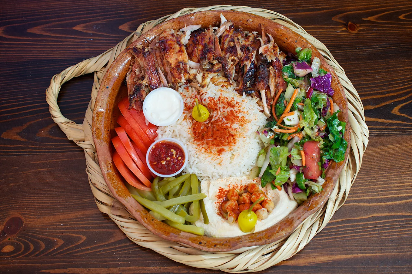 A large plate with grilled chicken, rice topped with spices, sliced tomatoes, green pickles, a green salad, hummus, a yellow pepper, a small cup of sauce, and dollops of yogurt, served on a wooden table.