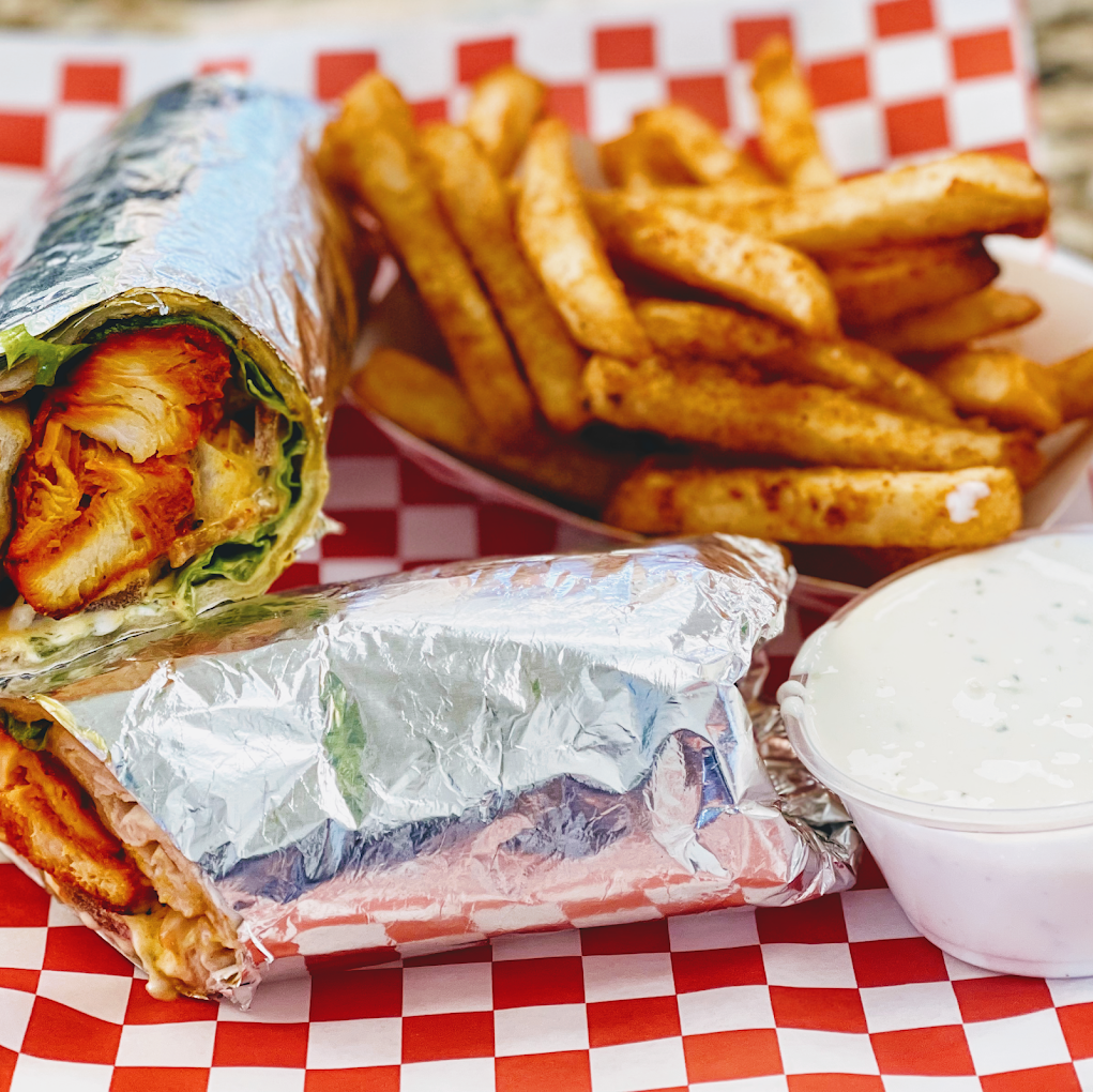 A foil-wrapped chicken wrap with lettuce, served with seasoned fries on red-checkered paper and a cup of ranch dipping sauce.