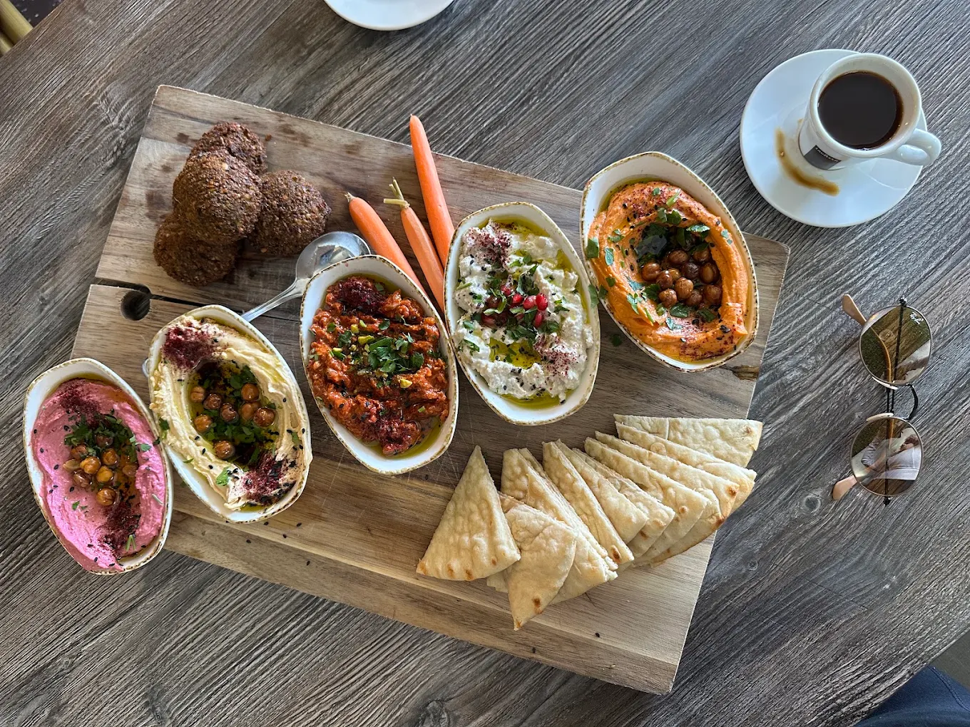 A wooden board with assorted dips, pita bread slices, falafel, carrot sticks, a spoon, and a cup of coffee on a rustic table. Sunglasses rest nearby.