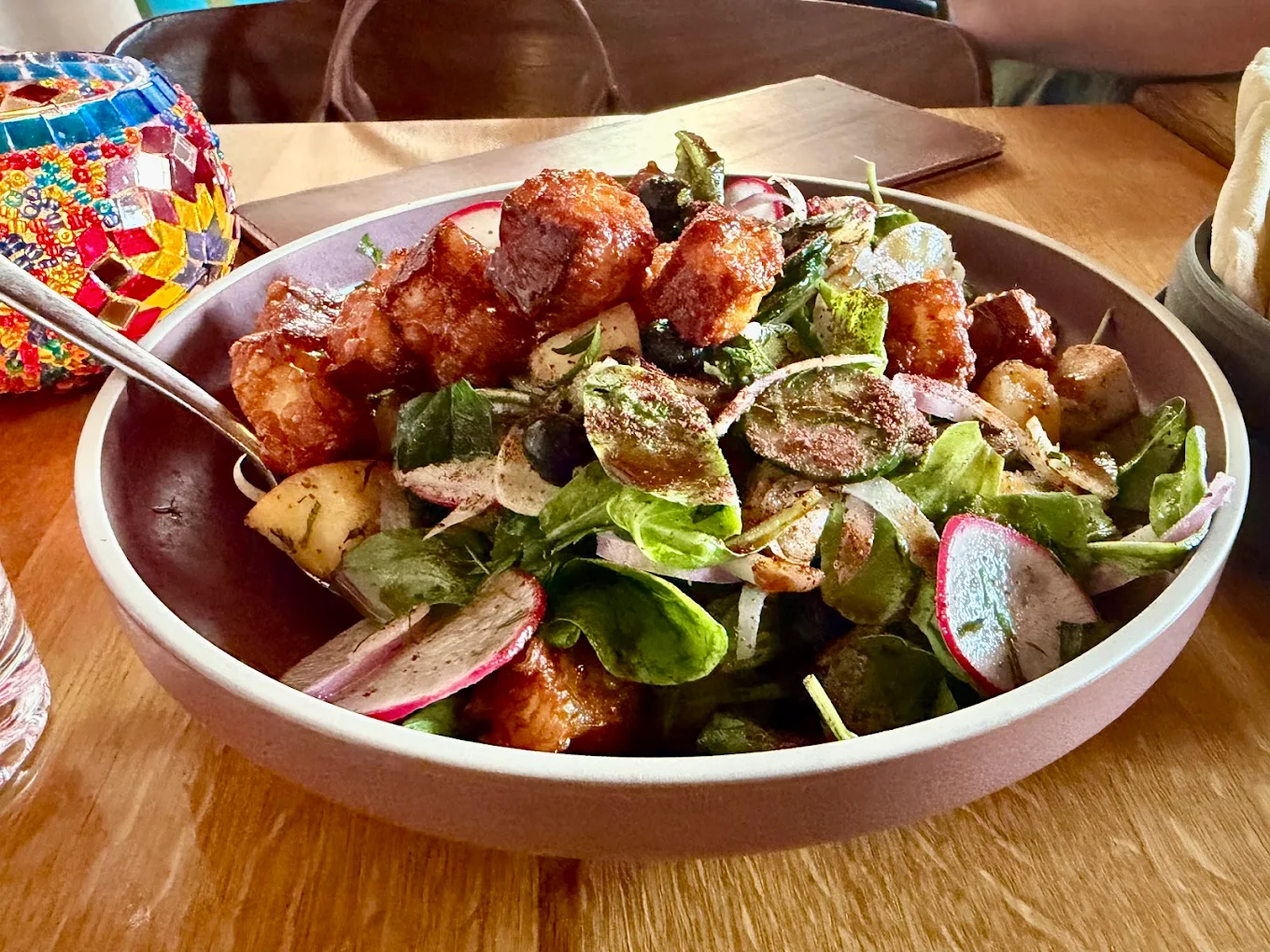 A bowl of fresh salad with greens, radishes, roasted potatoes, and crispy fried cubes, served with a fork on a wooden table next to a colorful mosaic lamp.