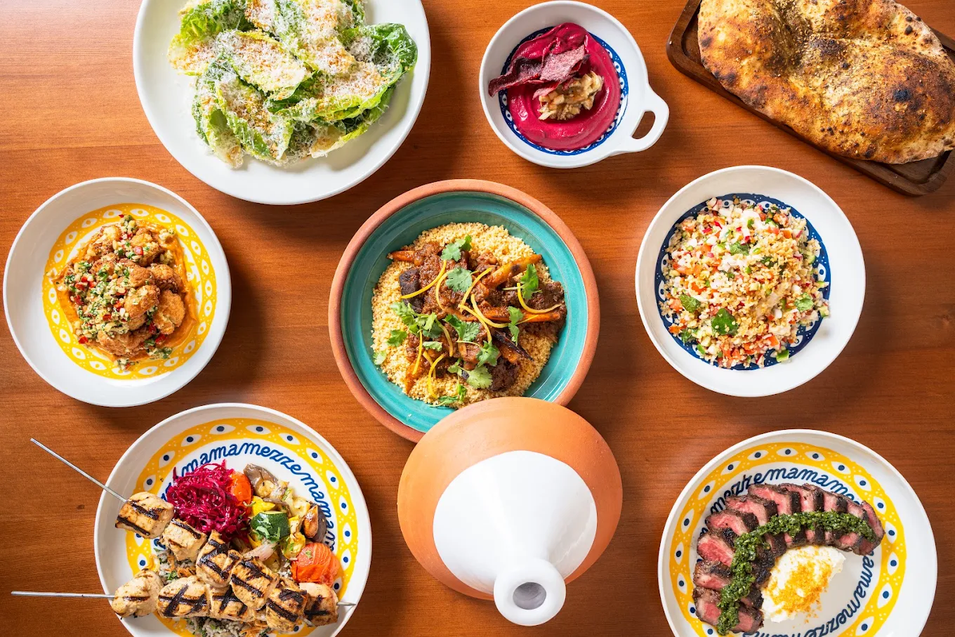 A table set with colorful Mediterranean dishes, including salad, bread, couscous, grilled skewers, vegetables, and garnish, all arranged in decorative bowls and plates.