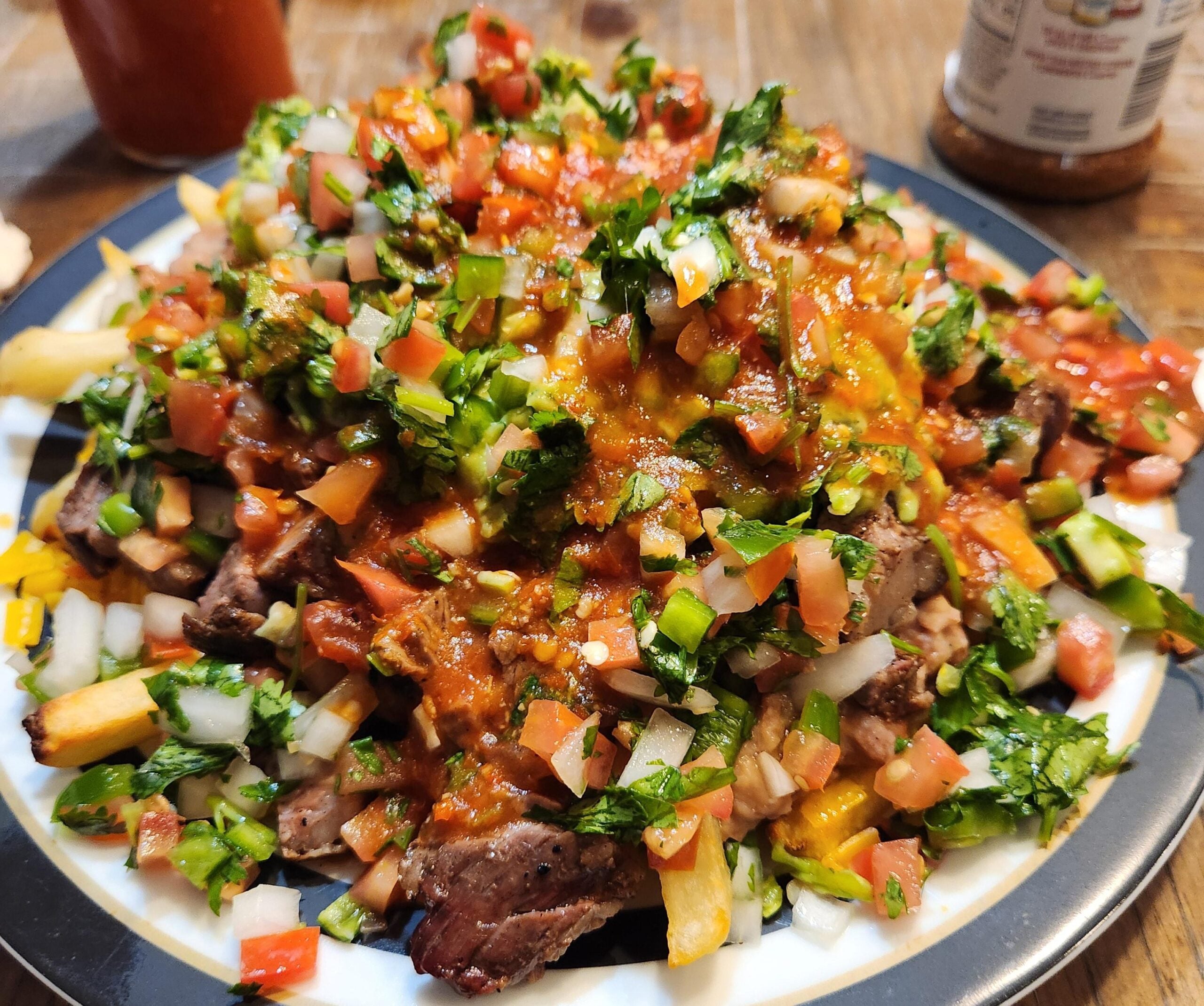 A plate of carne asada fries topped with melted cheese, chopped tomatoes, onions, cilantro, and sauce, served on a wooden table.