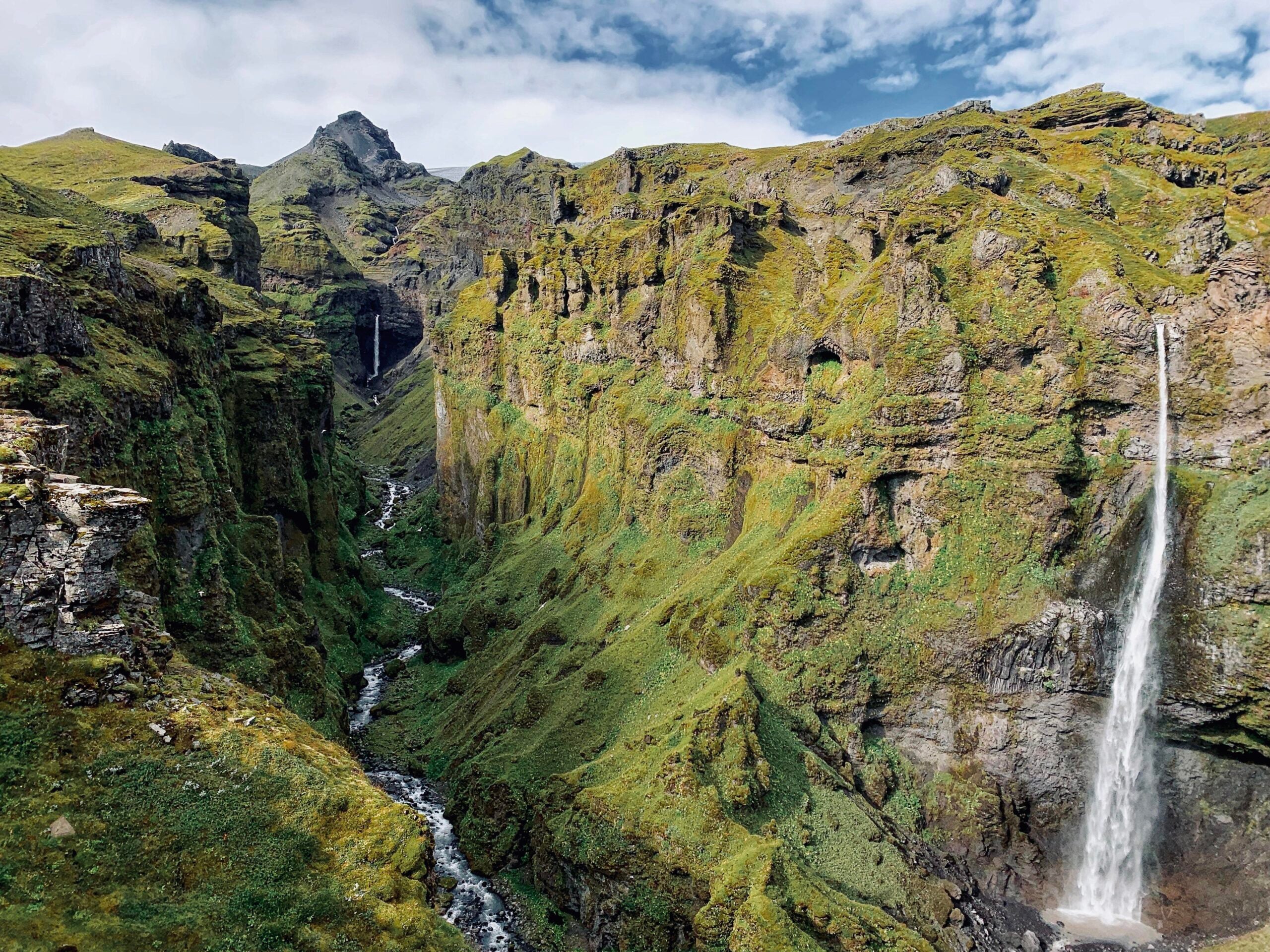 A lush green canyon with two tall, narrow waterfalls cascading down rocky cliffs under a partly cloudy sky. A small stream flows through the valley below.