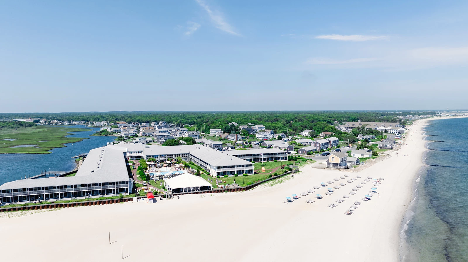Aerial view of a beachfront resort with white buildings, green lawns, and lounge chairs arranged along a sandy beach beside the ocean. Other resorts, houses, and lush greenery are visible in the background.