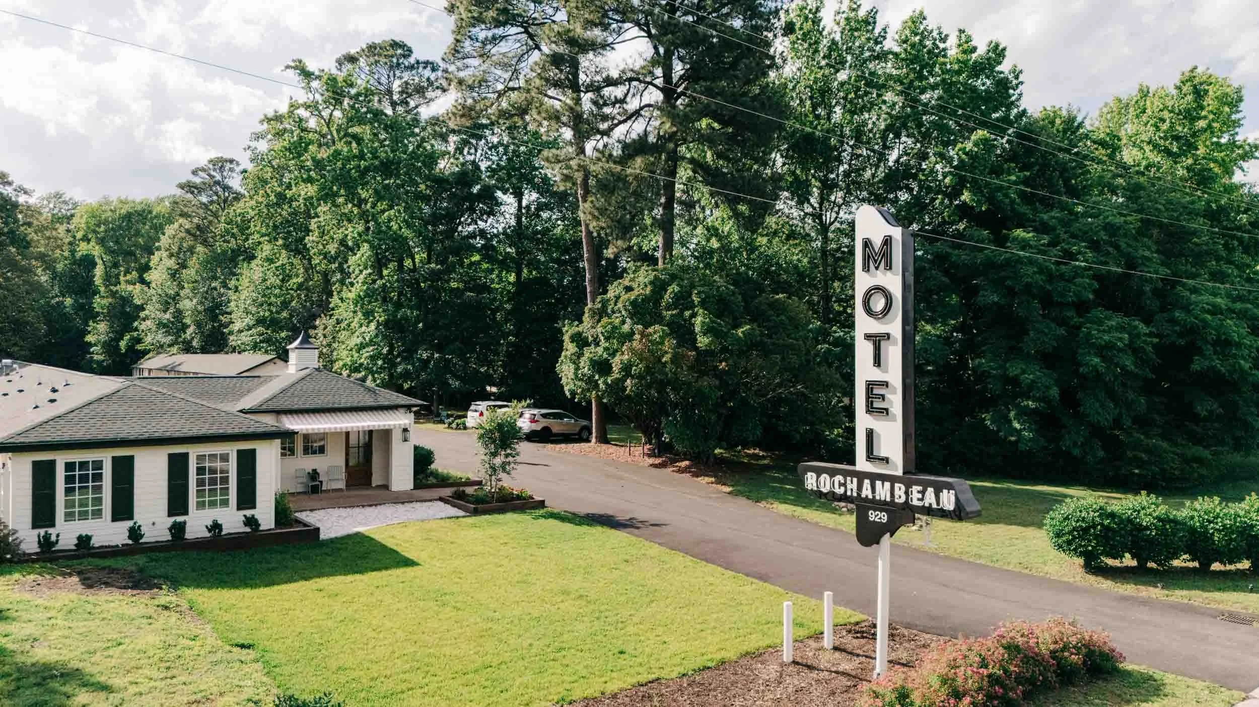 A small white motel with a vintage “MOTEL ROCHAMBEAU” sign stands by a driveway, surrounded by green grass, trees, and bushes on a sunny day—offering classic charm and the inviting atmosphere often found at resorts.