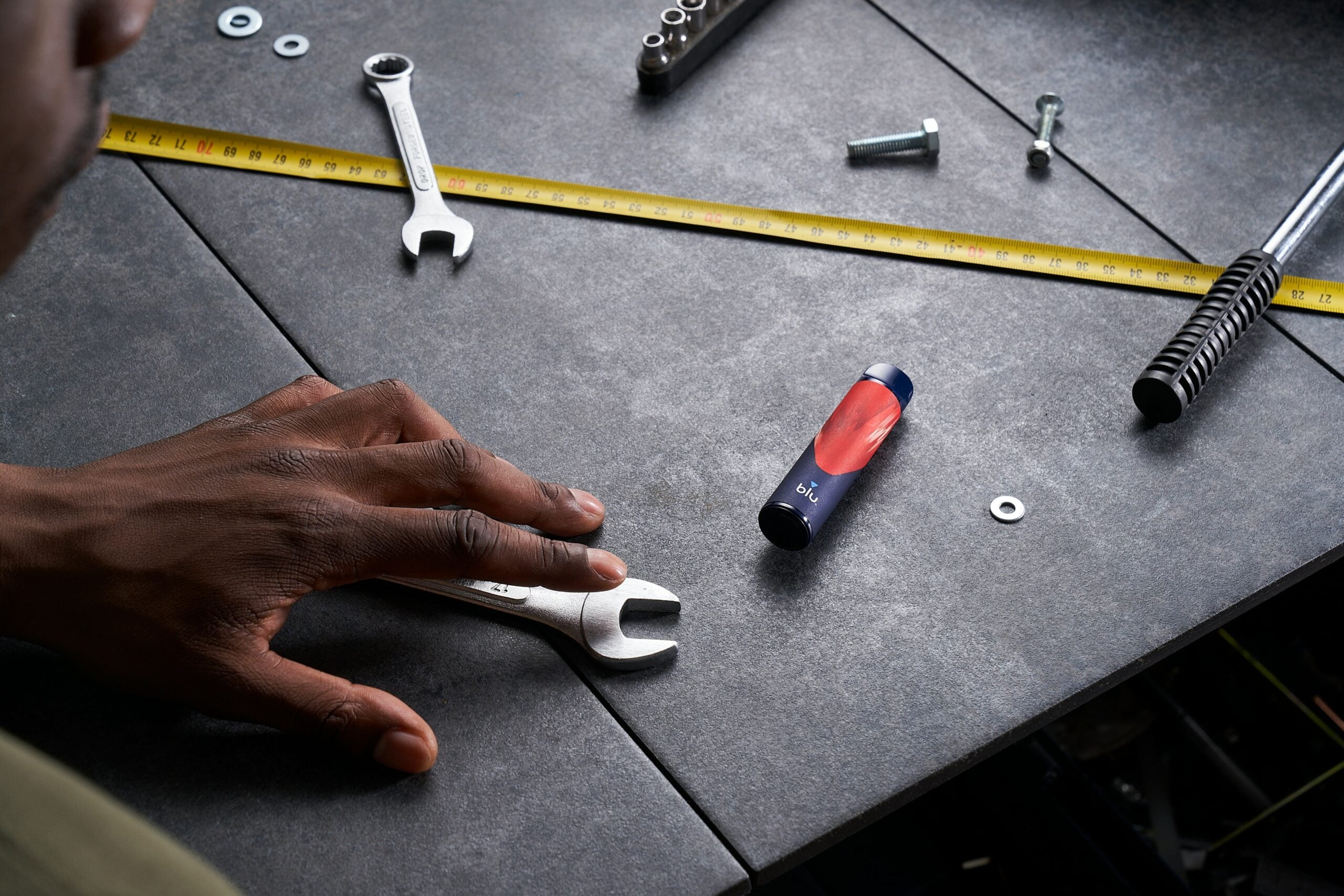 A hand holds a wrench on a gray workbench with a vape device, tape measure, bolt, screw, and other tools scattered around.