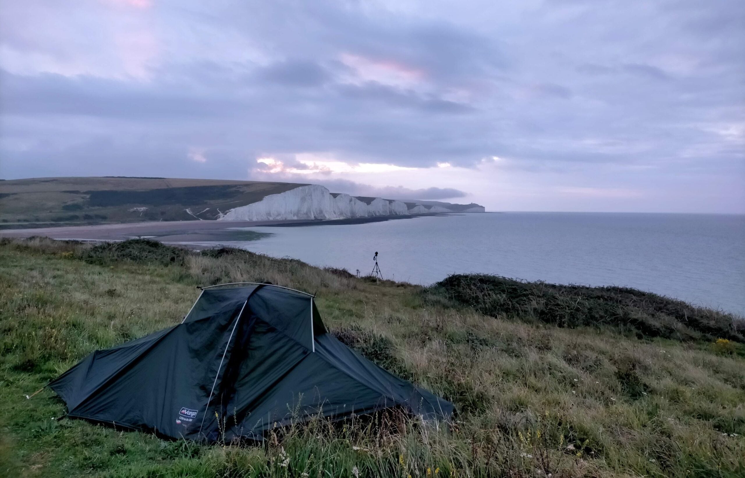 A green tent on grassy cliffs overlooks the sea and white chalk cliffs under a cloudy sky at dusk. A tripod stands nearby, capturing the scenic coastal landscape.