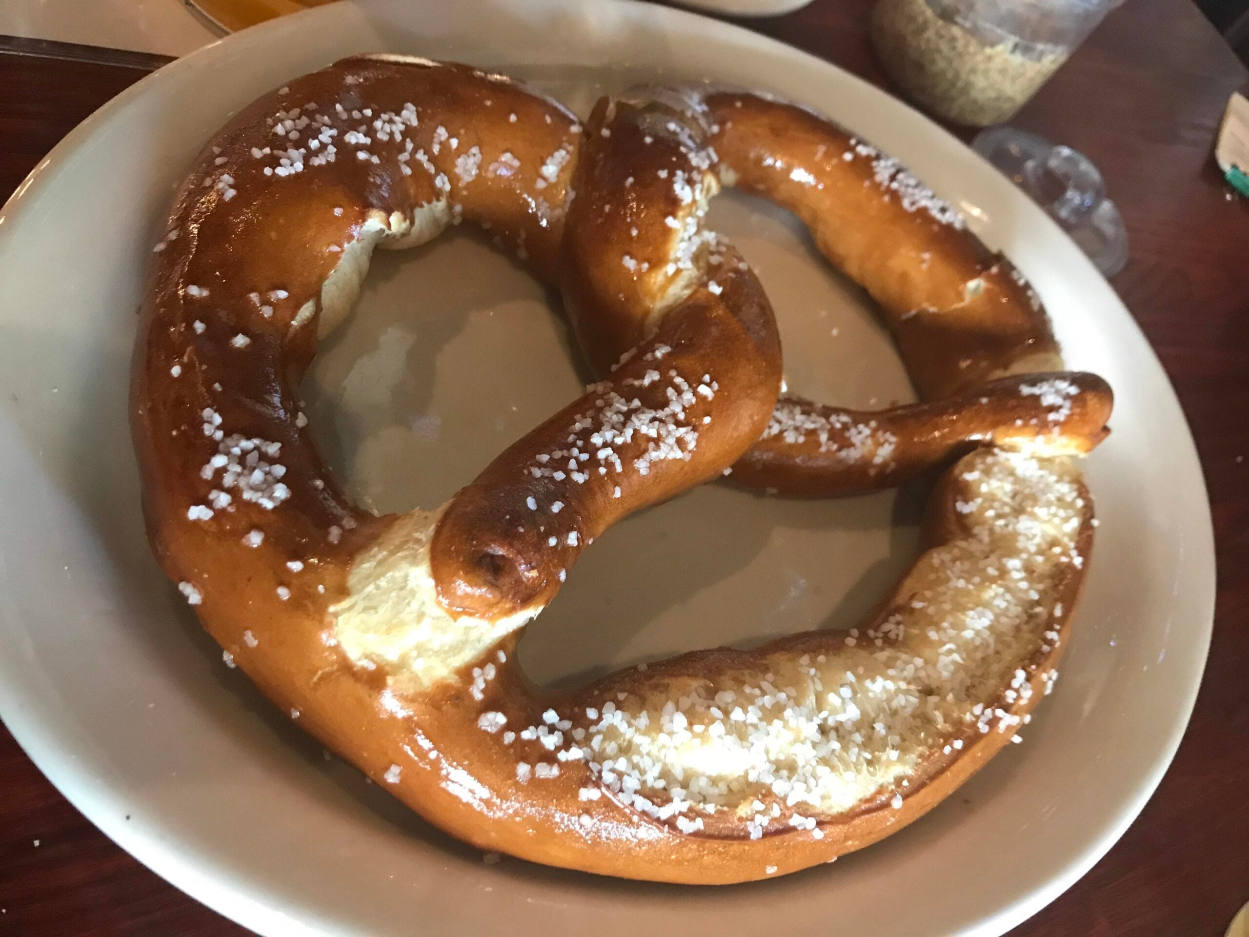 A large, golden-brown soft pretzel sprinkled with coarse salt sits on a white plate on a wooden table.