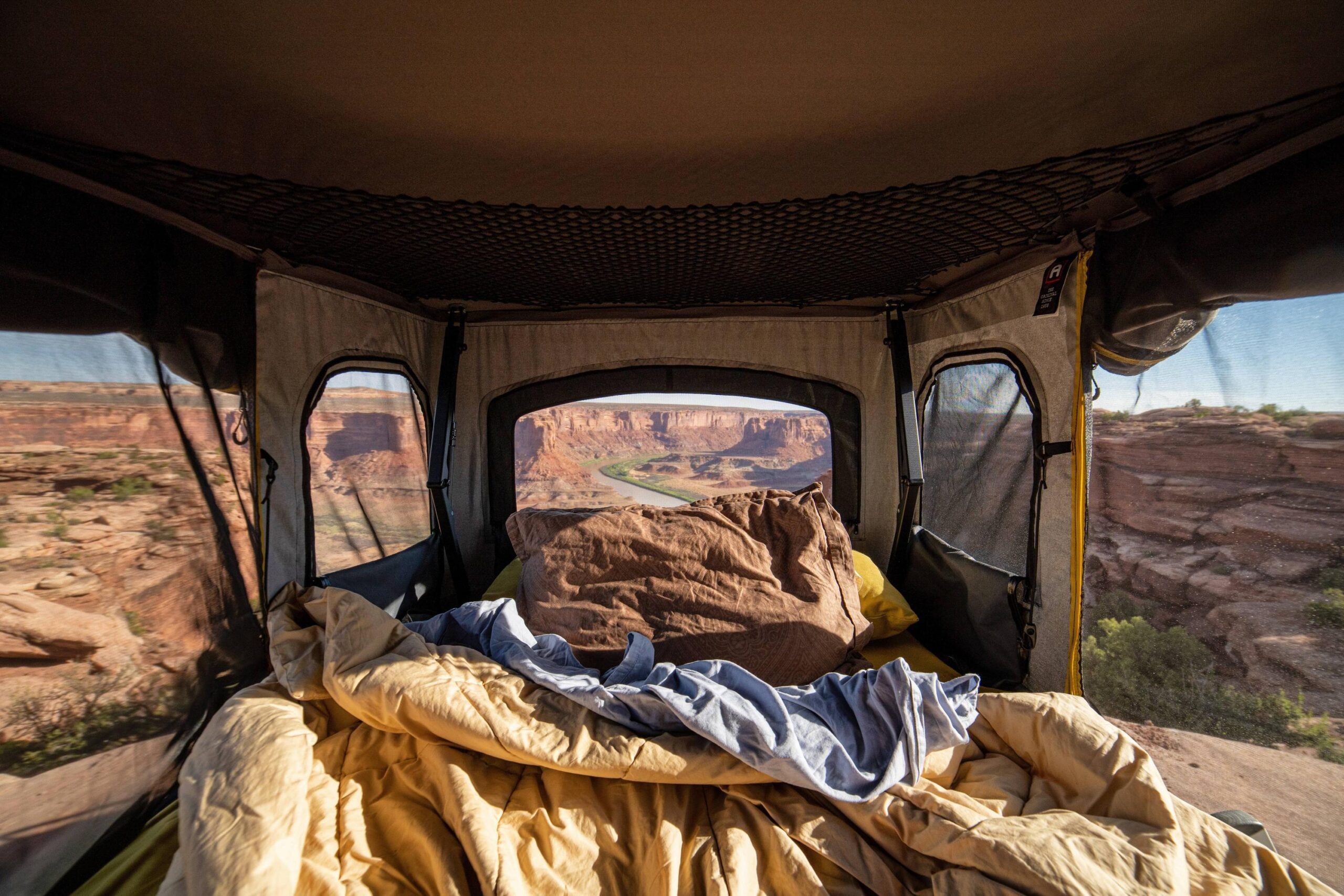 View from inside a tent, looking out over a canyon landscape with rocky cliffs and winding river. Bedding and pillows are spread out in the foreground.
