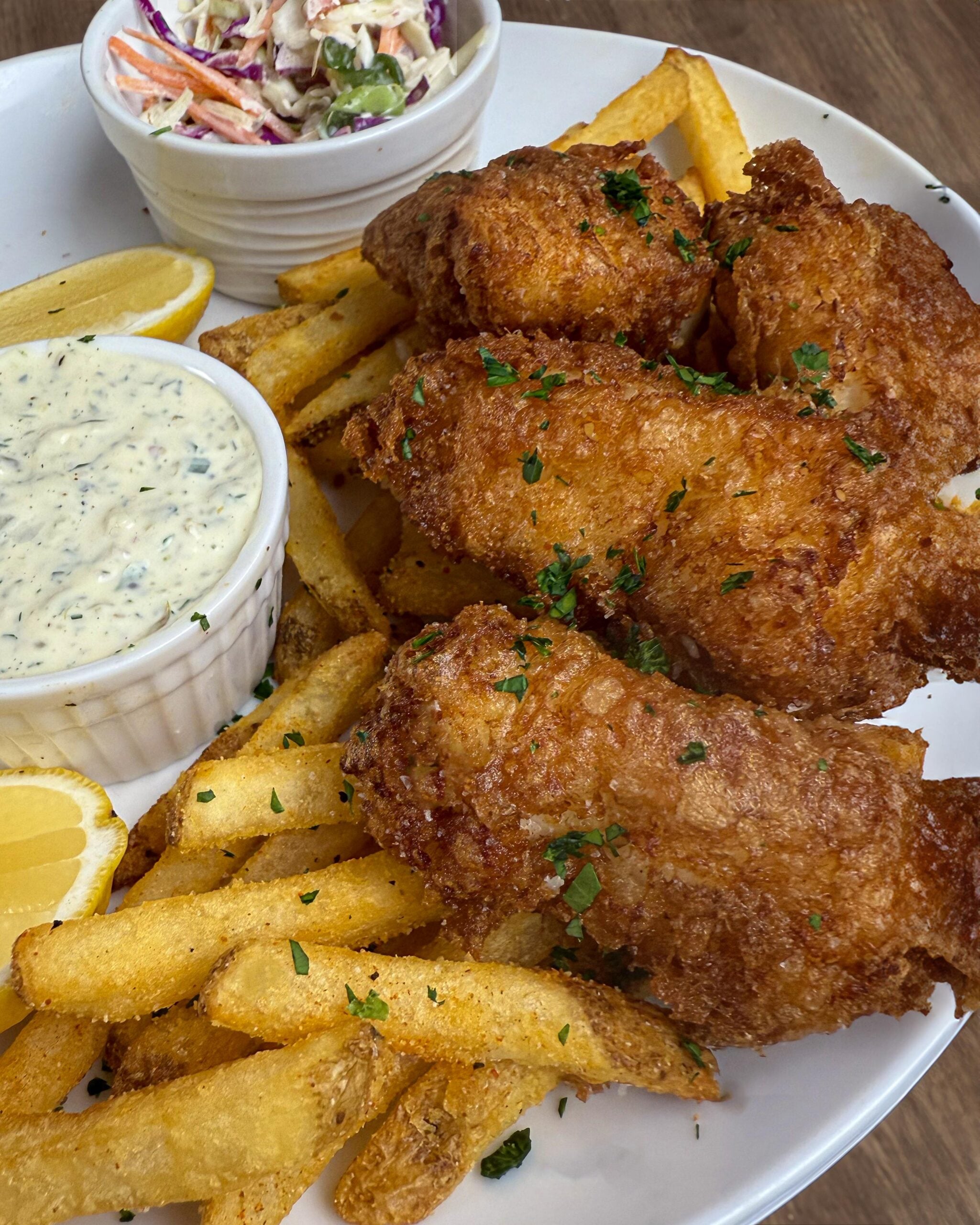 A plate of crispy fried fish, golden French fries, lemon wedges, tartar sauce, and a small bowl of coleslaw, garnished with herbs.