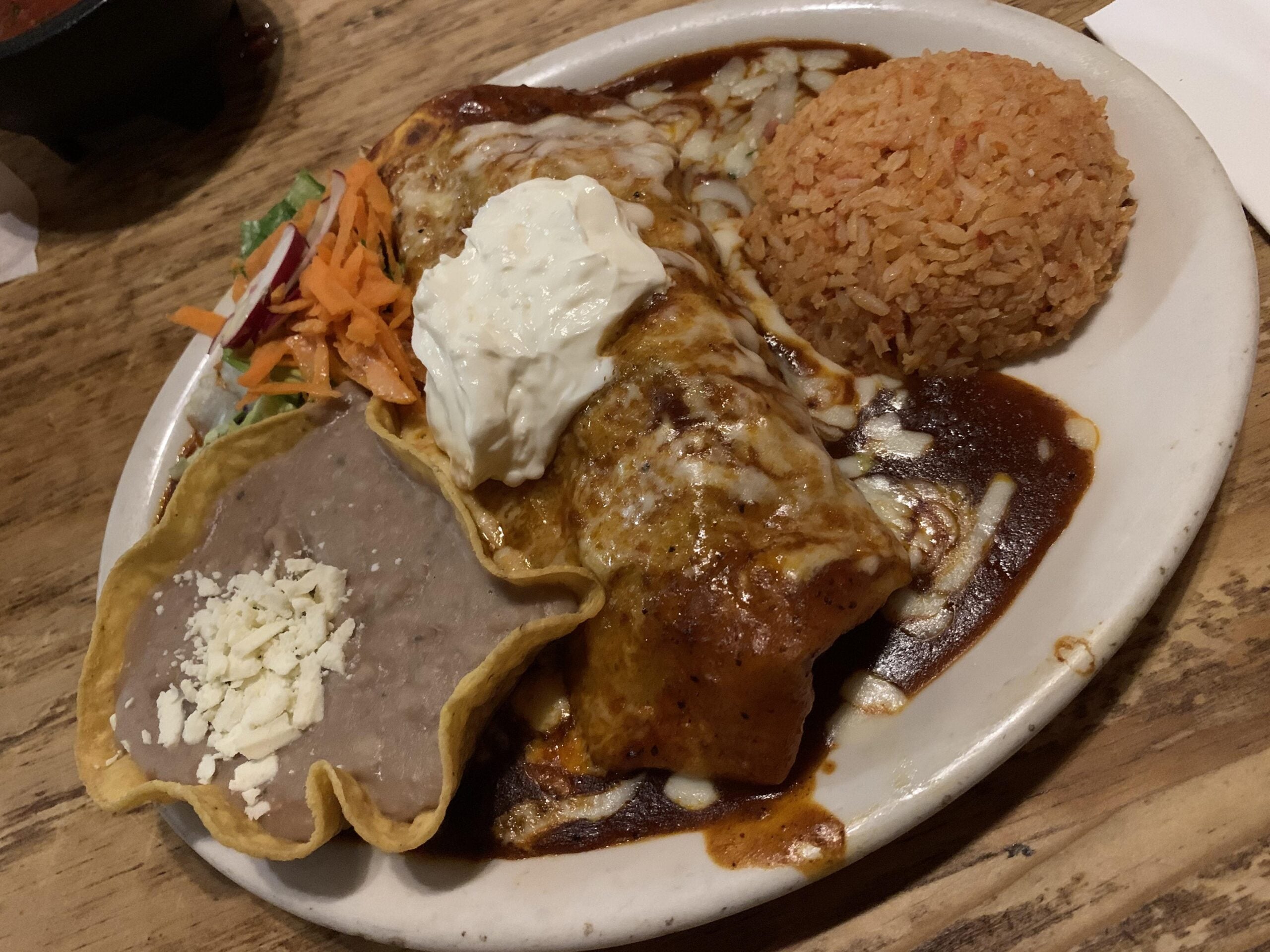 A plate with a saucy enchilada topped with sour cream, a mound of Mexican rice, refried beans in a tortilla shell with cheese, and a small side salad on a wooden table.