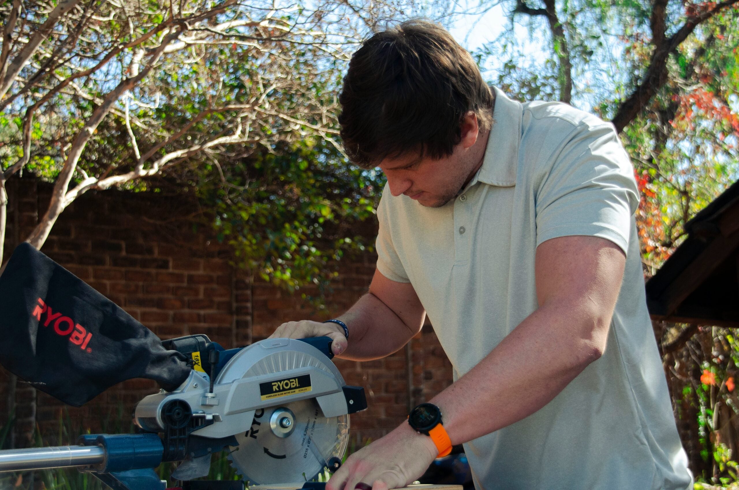 A person in a light polo shirt uses a Ryobi miter saw outdoors, surrounded by trees and a brick wall, focusing on their work.