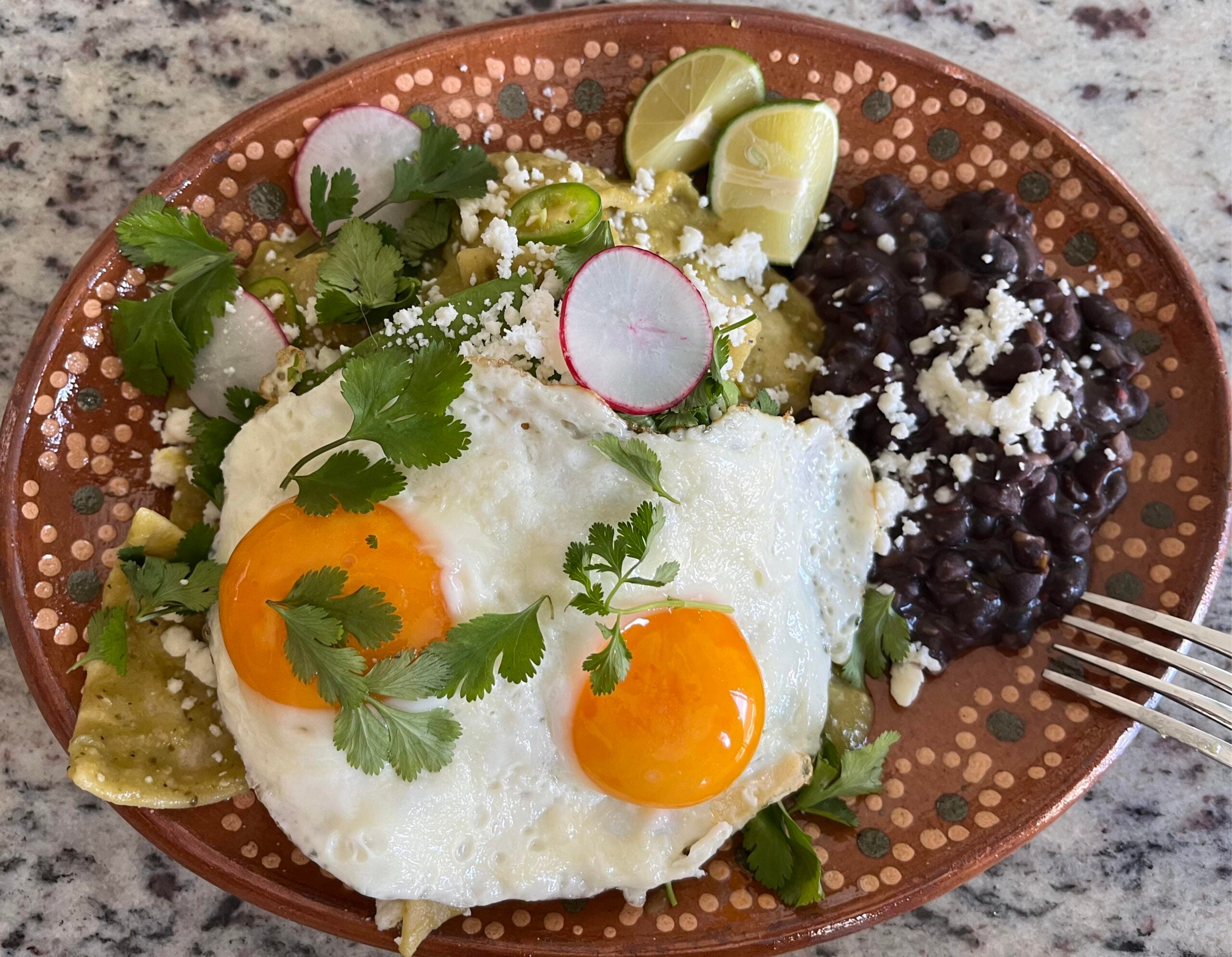 A plate of chilaquiles with green sauce, topped with two sunny-side-up eggs, cilantro, radishes, lime wedges, and a side of black beans with cheese. A fork rests on the plate.