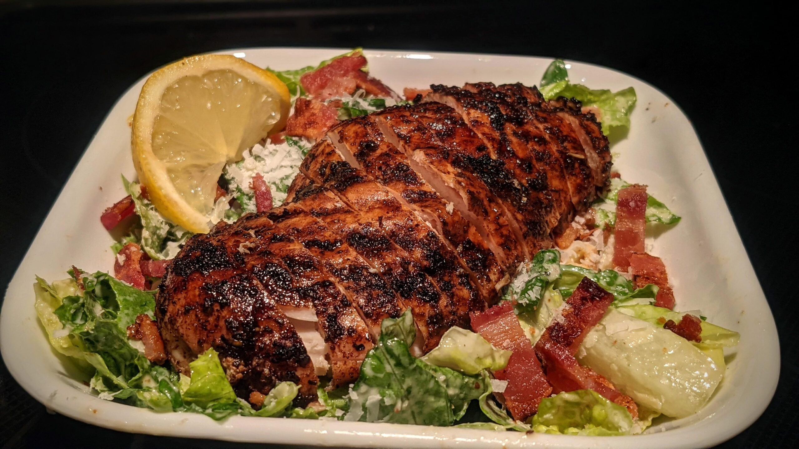 Grilled chicken breast on a bed of chopped salad with lettuce, bacon bits, shredded cheese, and a lemon wedge, served in a white dish.