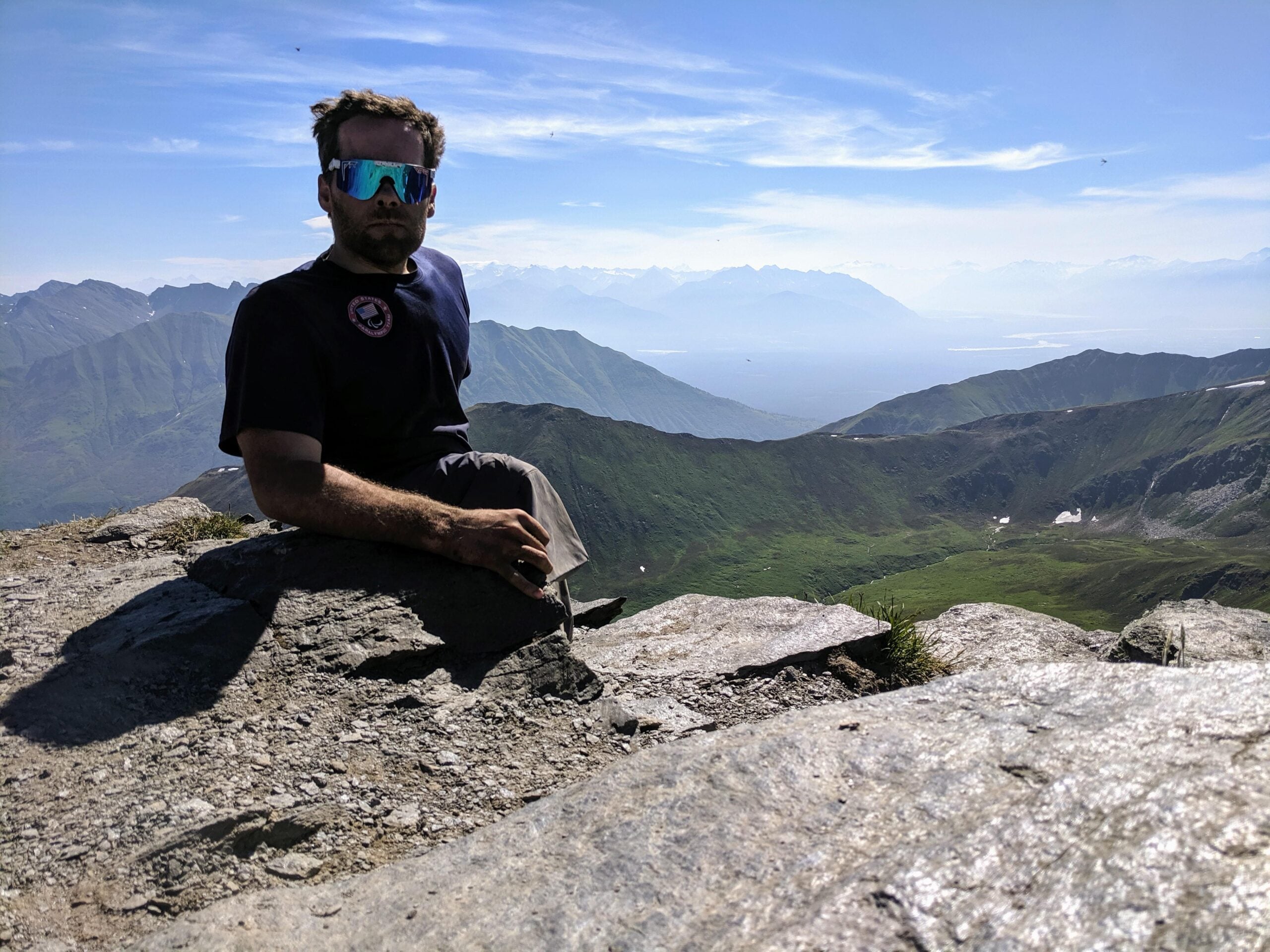 A man wearing sunglasses sits on a rocky mountain summit, posing for photos with green valleys and distant mountain ranges visible under a clear blue sky—a scene often posted by hikers seeking adventure.