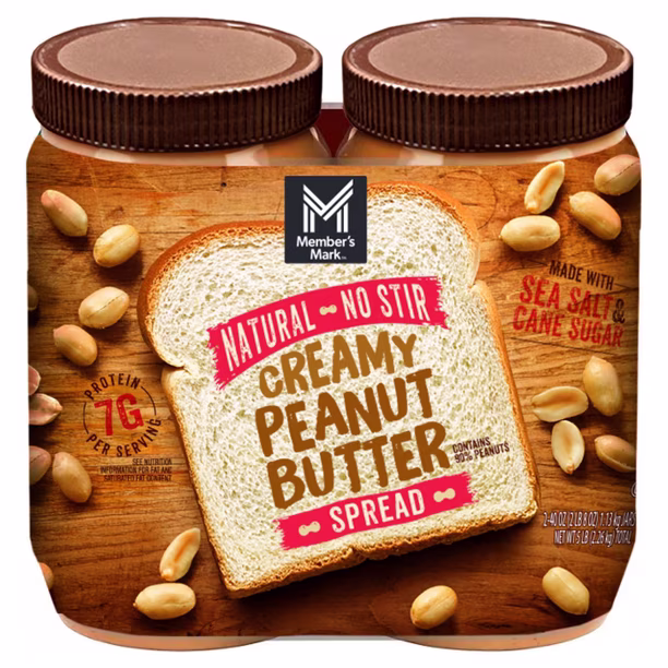 Two jars of Member’s Mark Creamy Peanut Butter Spread with brown lids, featuring an image of bread with peanut butter and scattered peanuts on the label. Text highlights “Natural,” “No Stir,” and “Made with sea salt & cane sugar.”