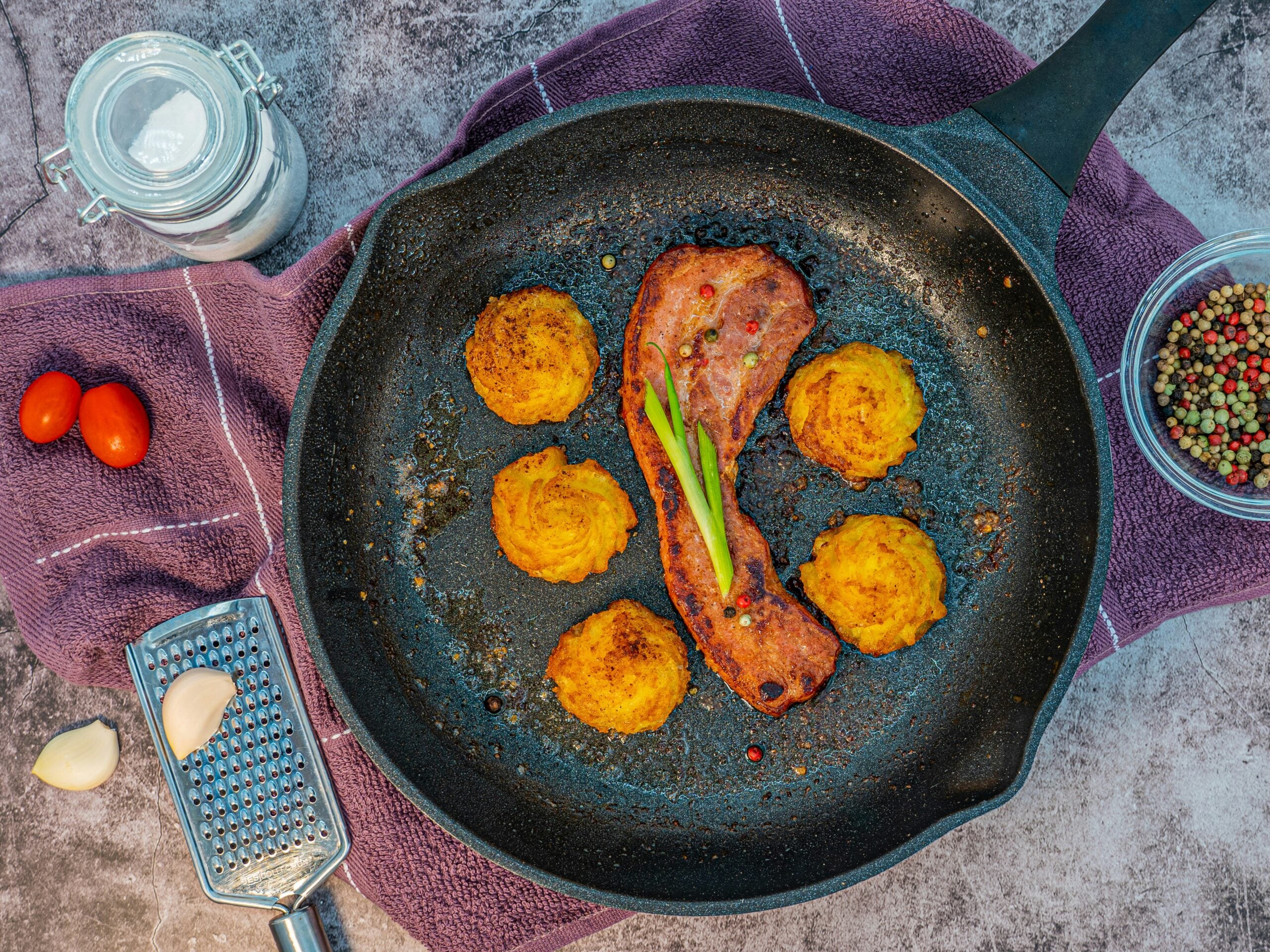 A frying pan with one strip of cooked bacon, six round potato croquettes, and green garnish. Surrounding the pan are cherry tomatoes, a jar of salt, garlic, a grater, and a bowl of peppercorns on a purple towel.