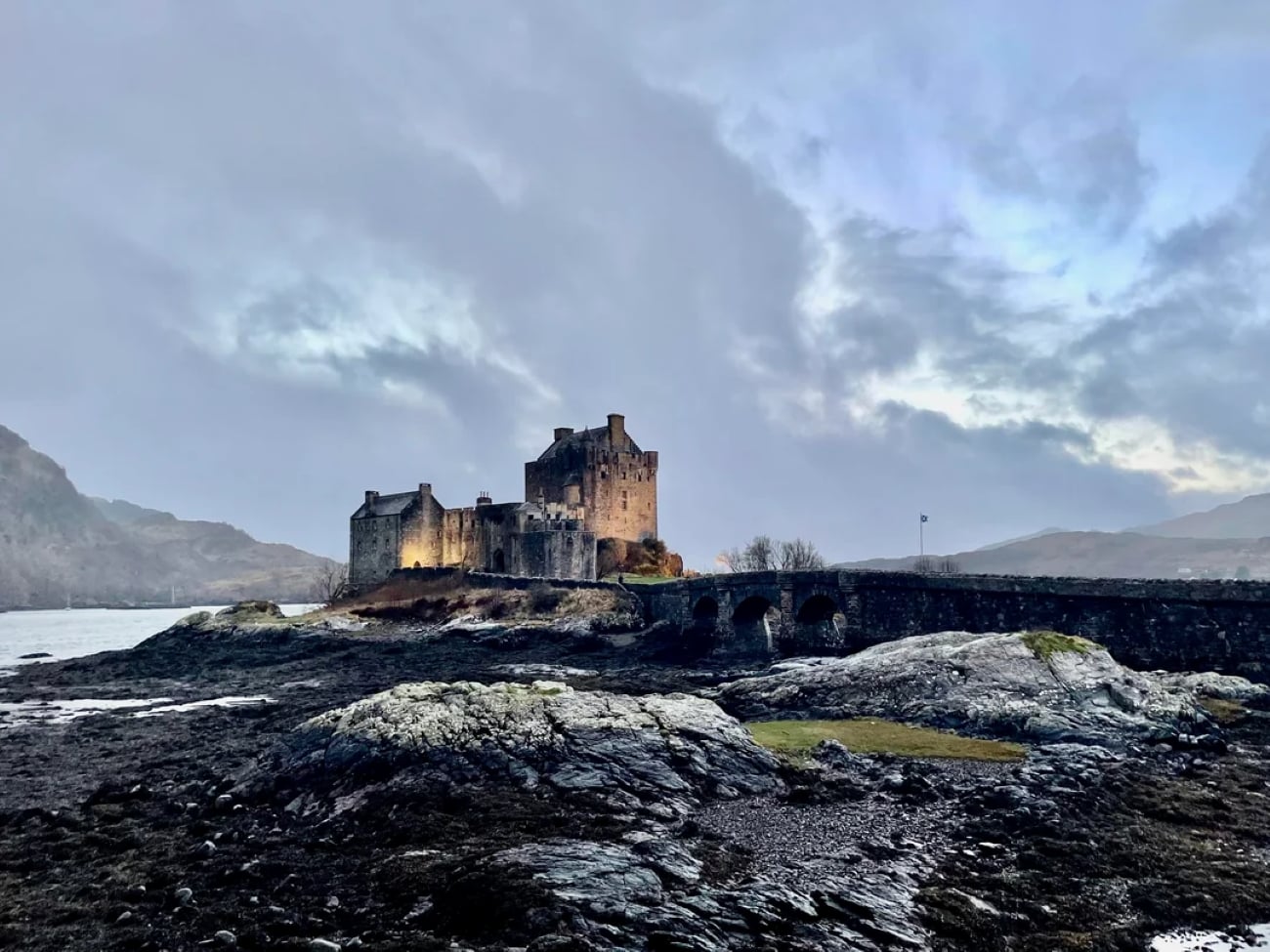 An old stone castle sits on an island, connected to land by a stone bridge, surrounded by rocky shores and water under a cloudy evening sky.