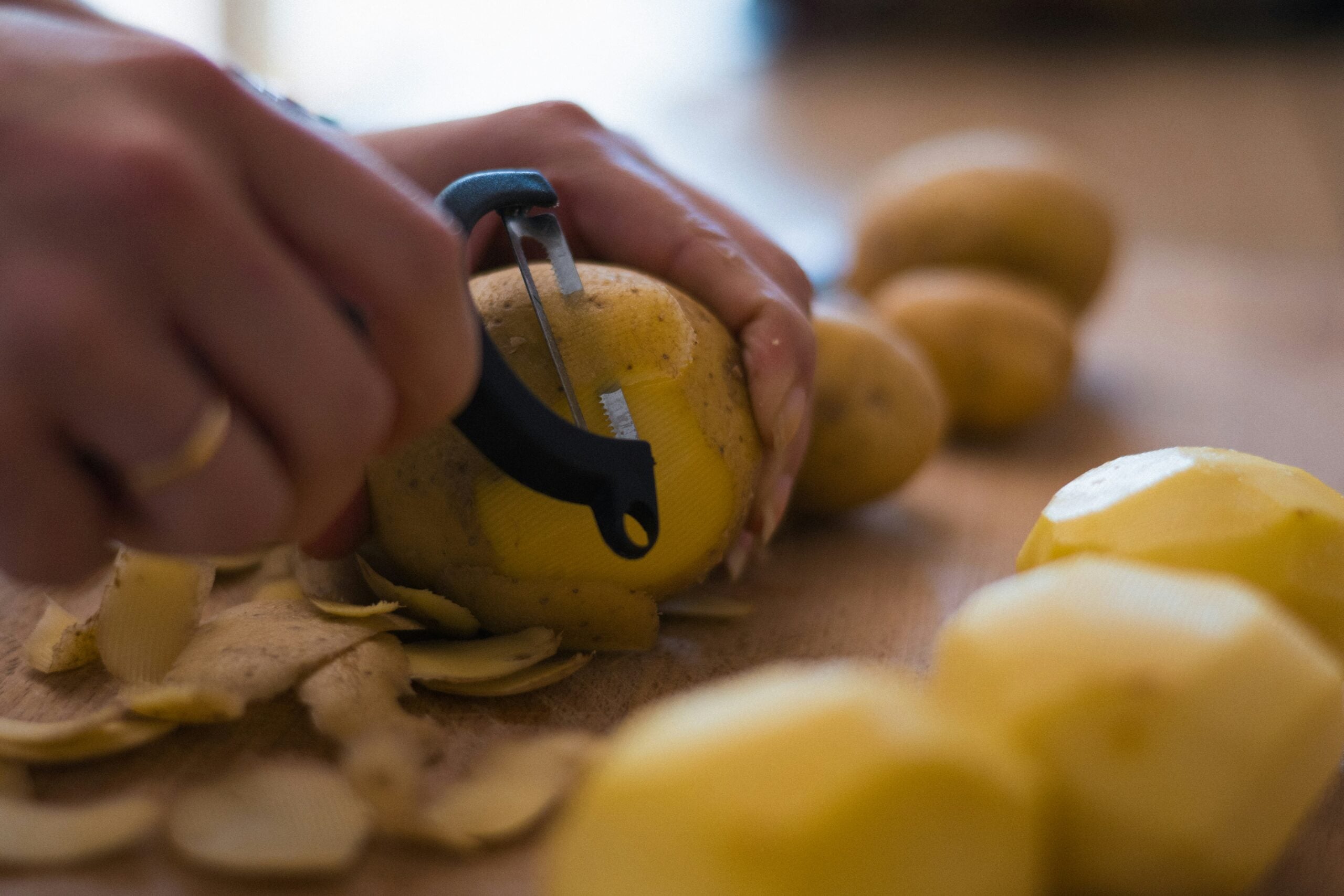 A person uses a black peeler to remove the skin from a potato on a wooden surface. Peeled potatoes and potato skins are also visible.