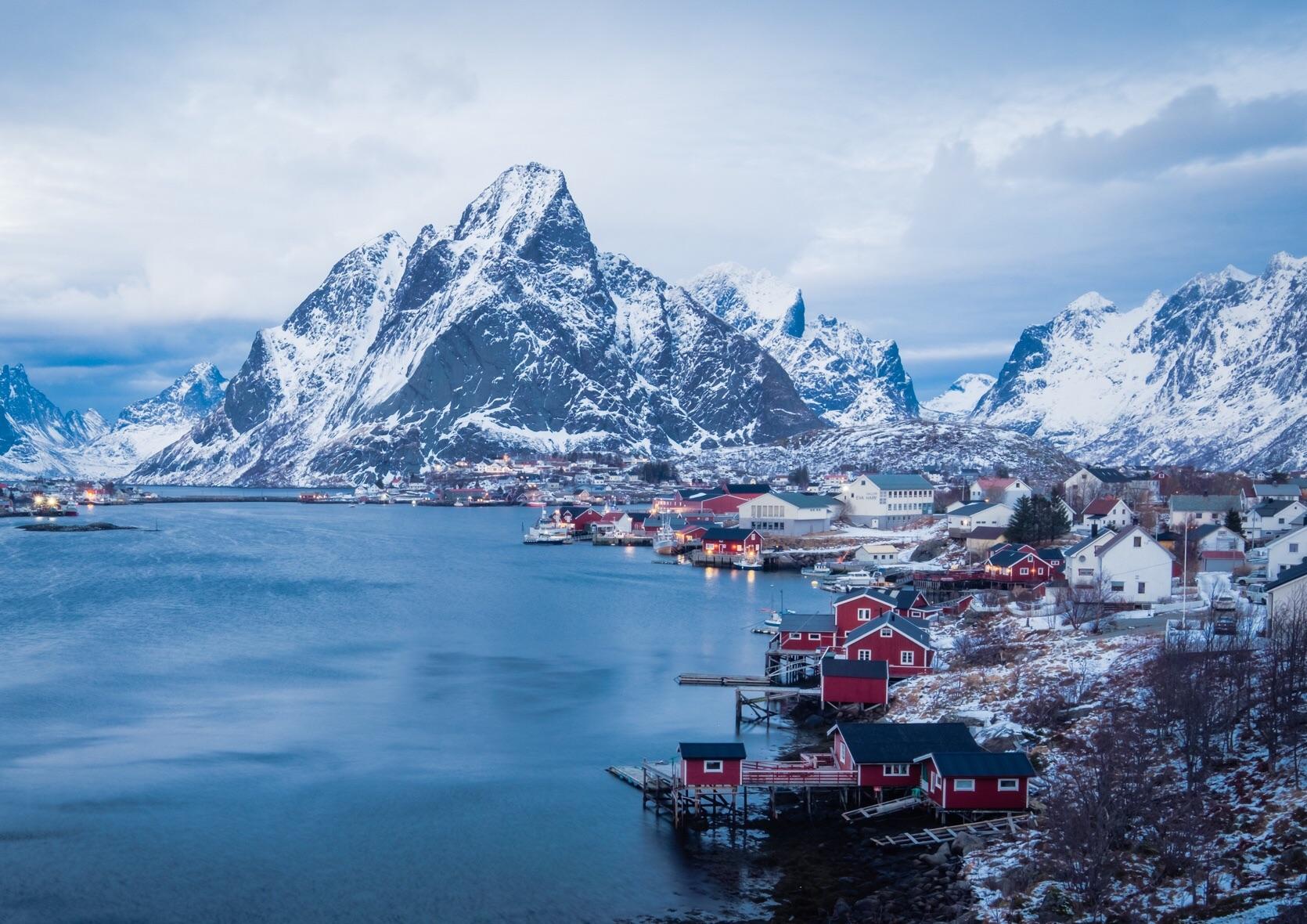 A snowy mountain towers over a fjord with red and white houses along the shore. The scene is peaceful with calm blue water, overcast sky, and winter landscape.