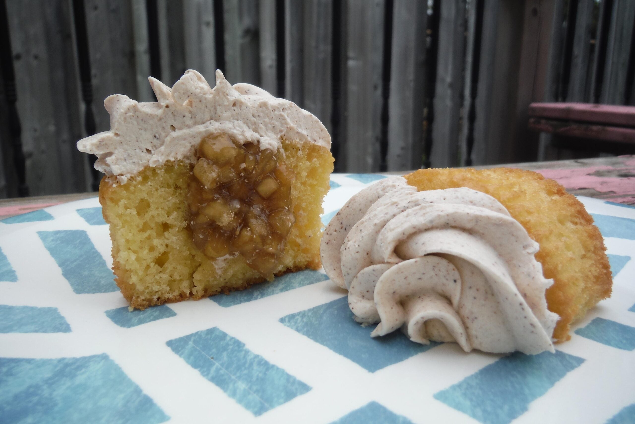 A sliced cupcake showing a spiced apple filling and topped with cinnamon frosting sits on a blue and white patterned plate outdoors.