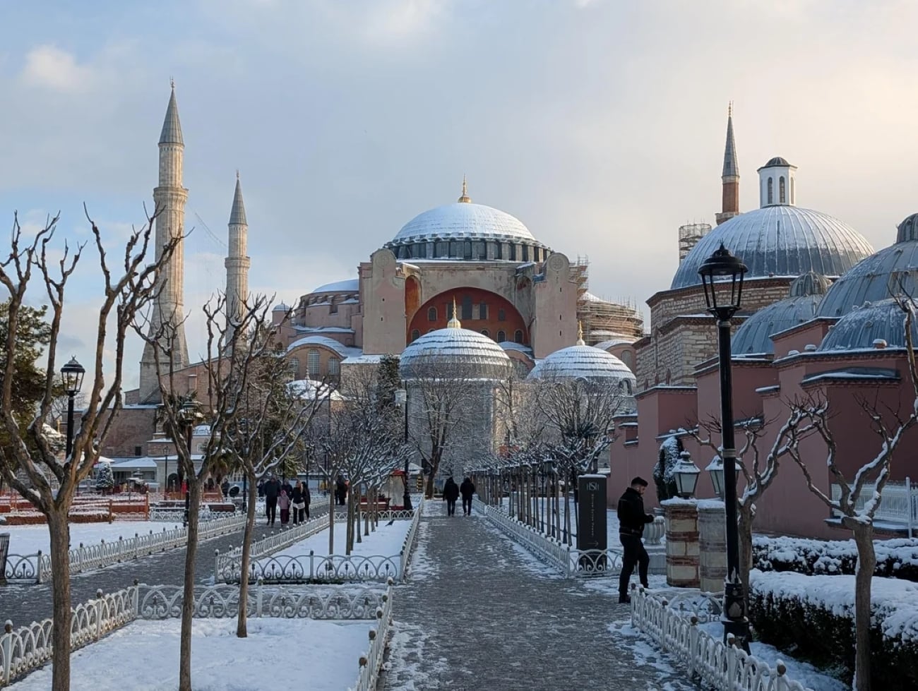 A snowy walkway leads to the Hagia Sophia in Istanbul, with bare trees and people walking along the path. The domes and minarets are dusted with snow under a partly cloudy sky.