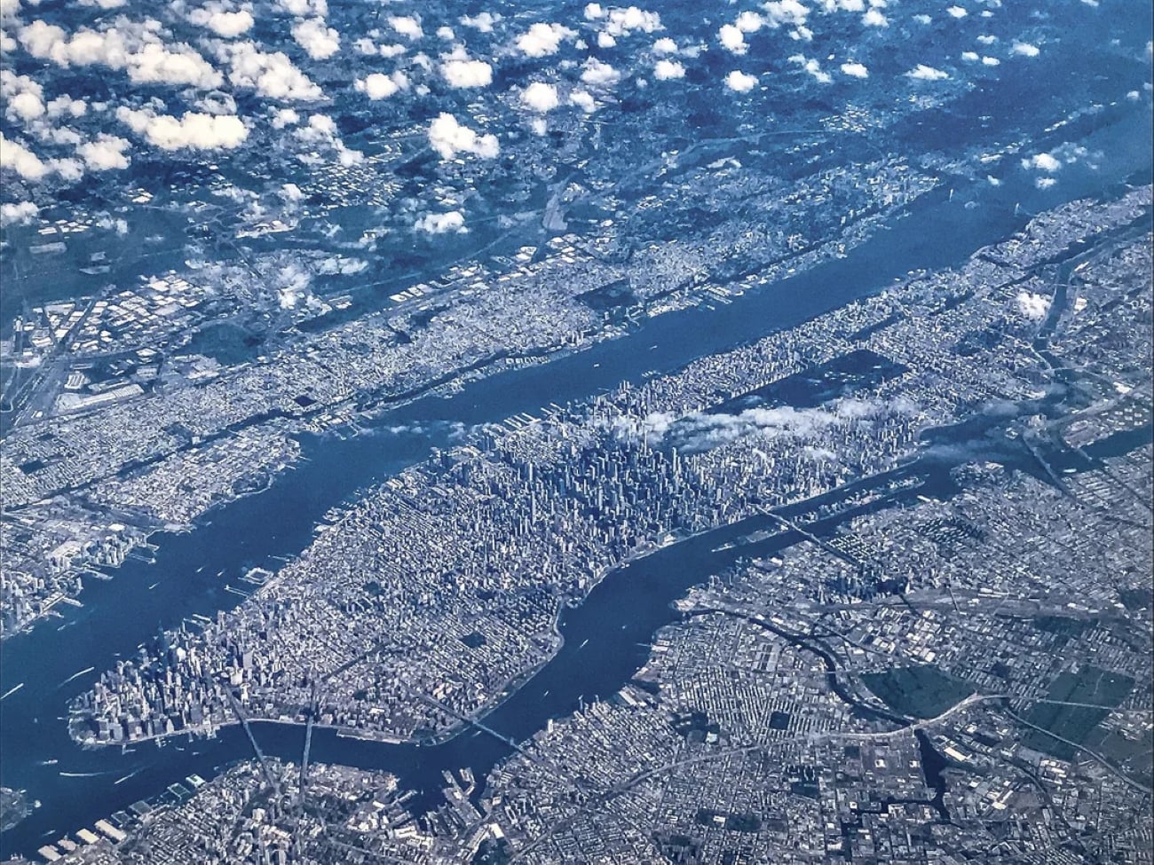 Aerial view of Manhattan, New York City, as seen through airplane windows, showing dense skyscrapers, surrounding rivers, and patches of clouds casting shadows over the urban landscape.