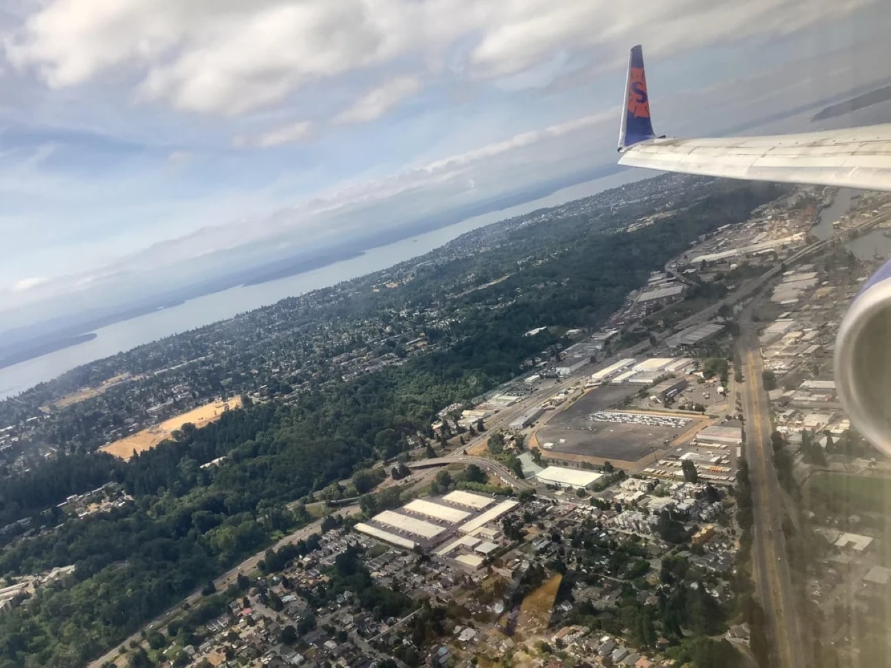 Aerial view of a city and greenery as seen through airplane windows, with part of the plane’s wing visible against a cloudy sky.