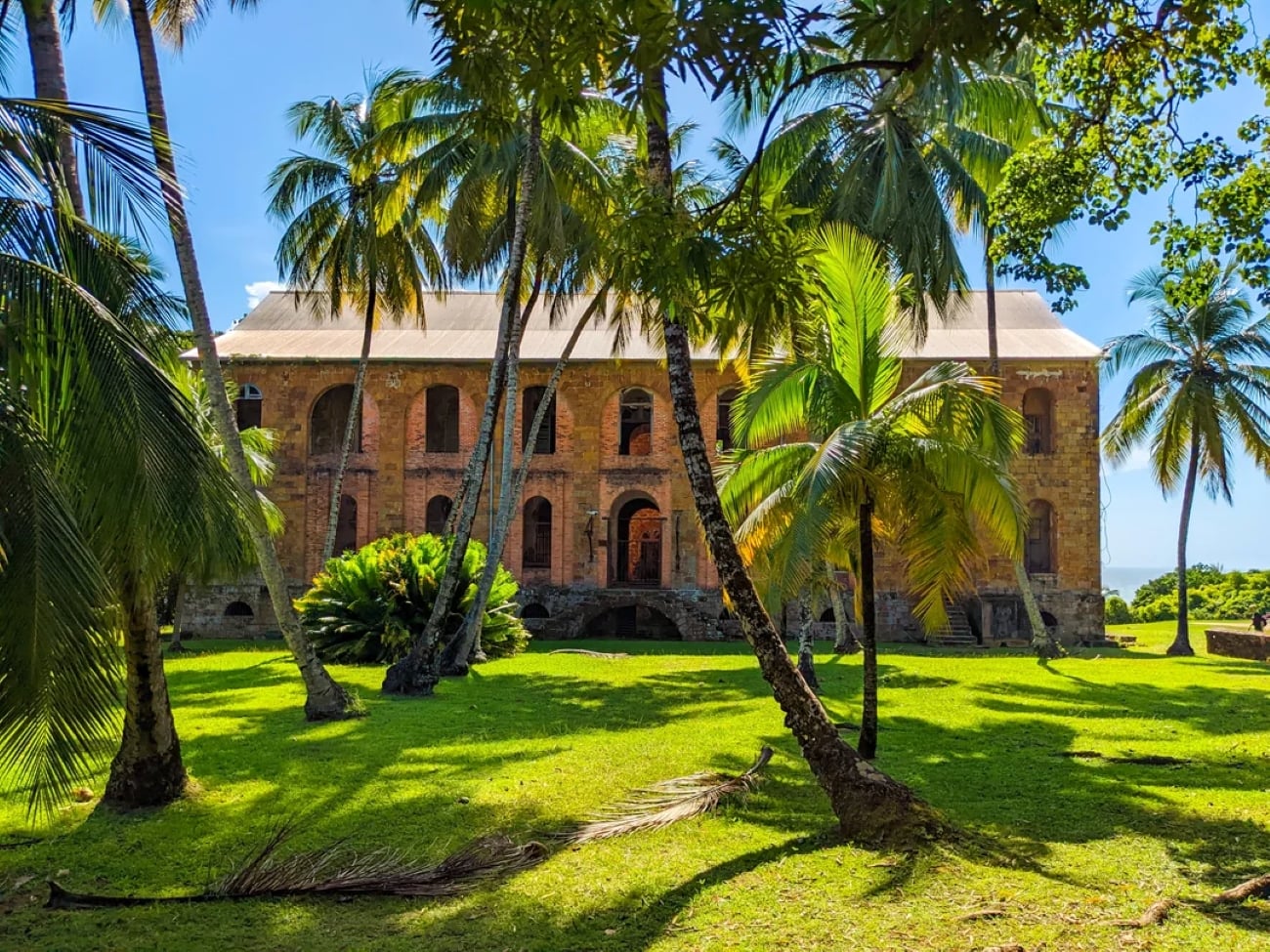 A historic brick building with arched windows stands among tall palm trees on a bright, sunny day. The lush green lawn and blue sky create a serene atmosphere reminiscent of the world’s most beautiful tropical locations.
