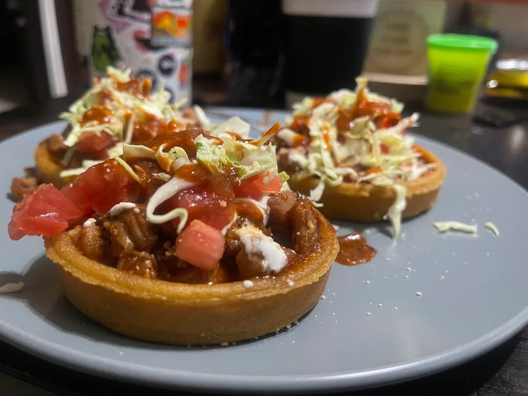 Three small savory tart shells filled with meat, chopped tomatoes, shredded lettuce, crumbled cheese, and drizzled with red sauce, served on a gray plate with blurred background items.